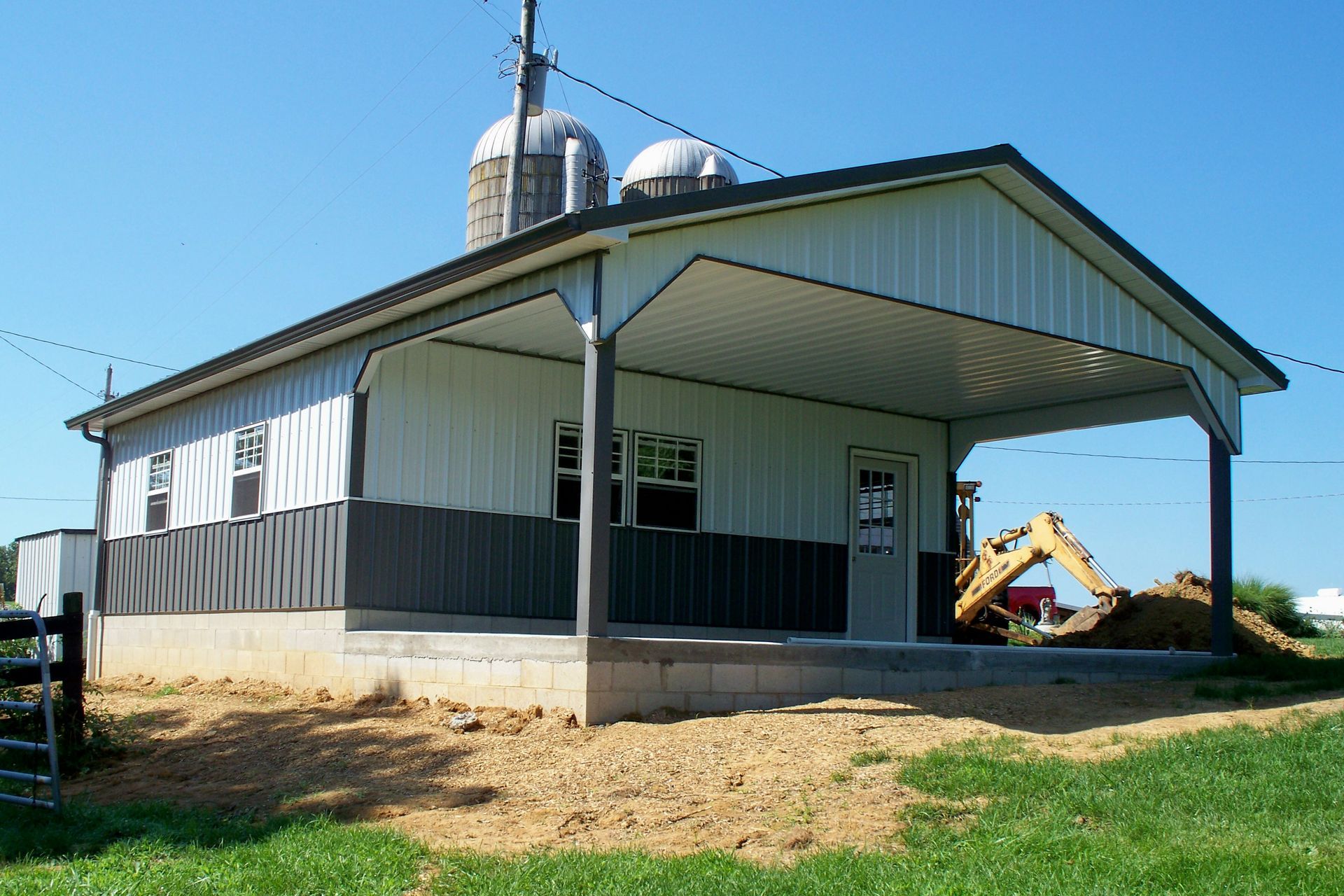 Farm building with metal roof and silo, a yellow excavator in the background, on a grassy lot.