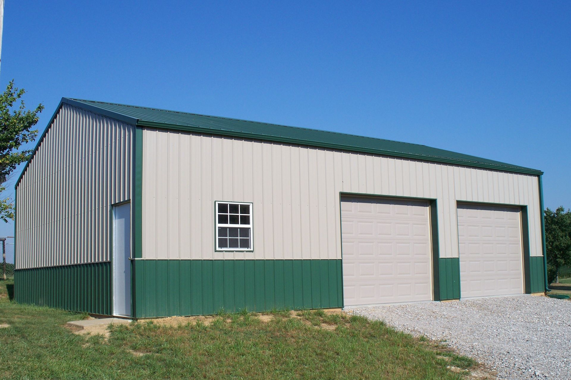 Green and white metal warehouse with two garage doors and a gravel driveway under a blue sky