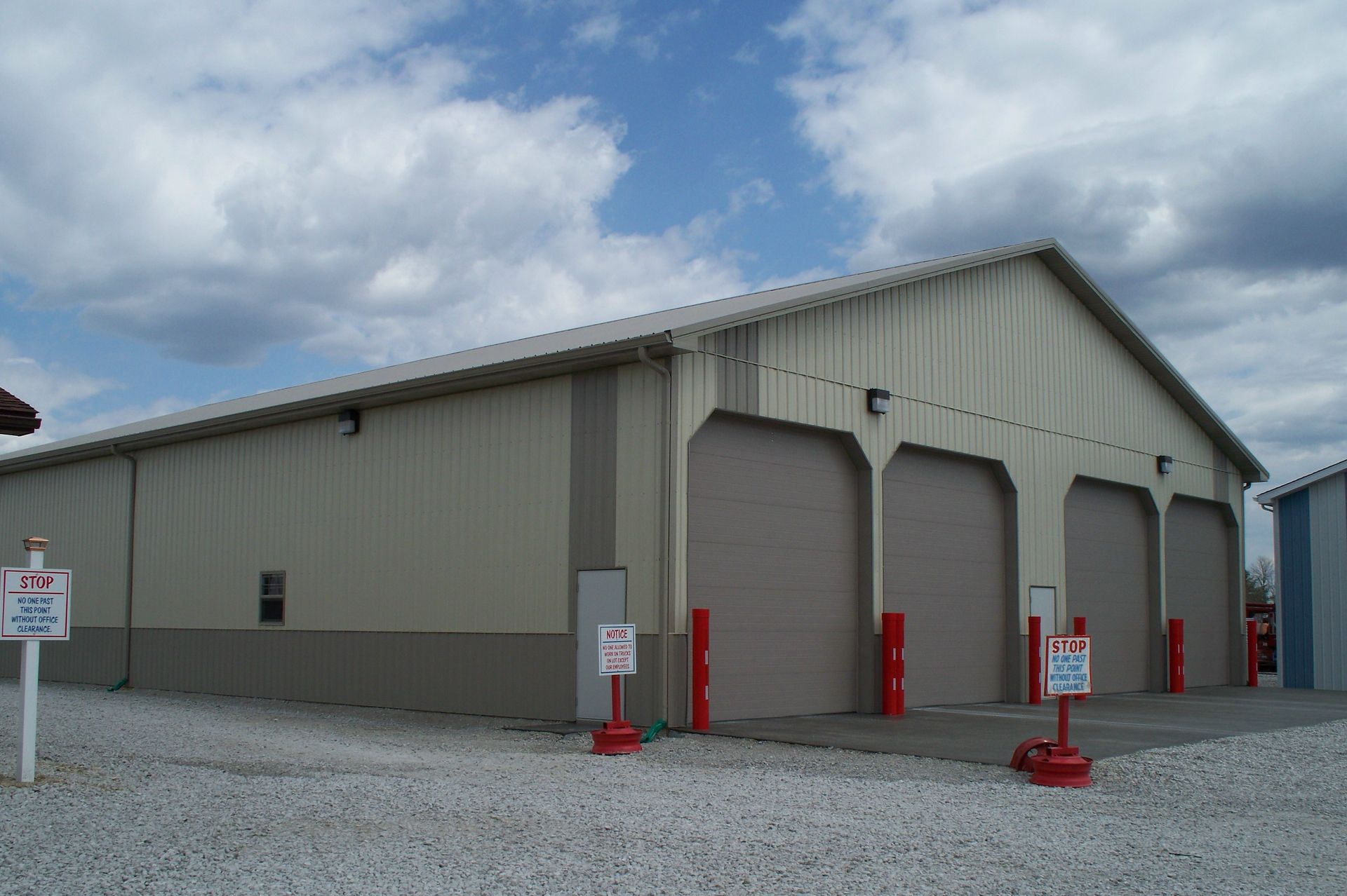 Large gray metal warehouse with three garage bays and red bollards under a cloudy sky