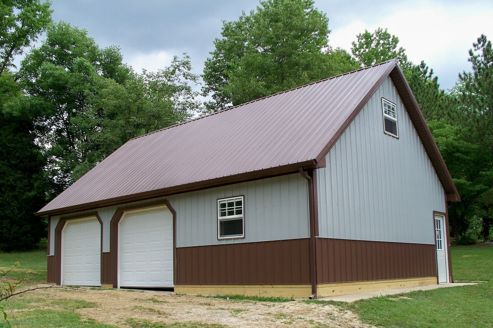 Brown and white barn with a gambrel roof and two garage doors in a grassy, wooded area