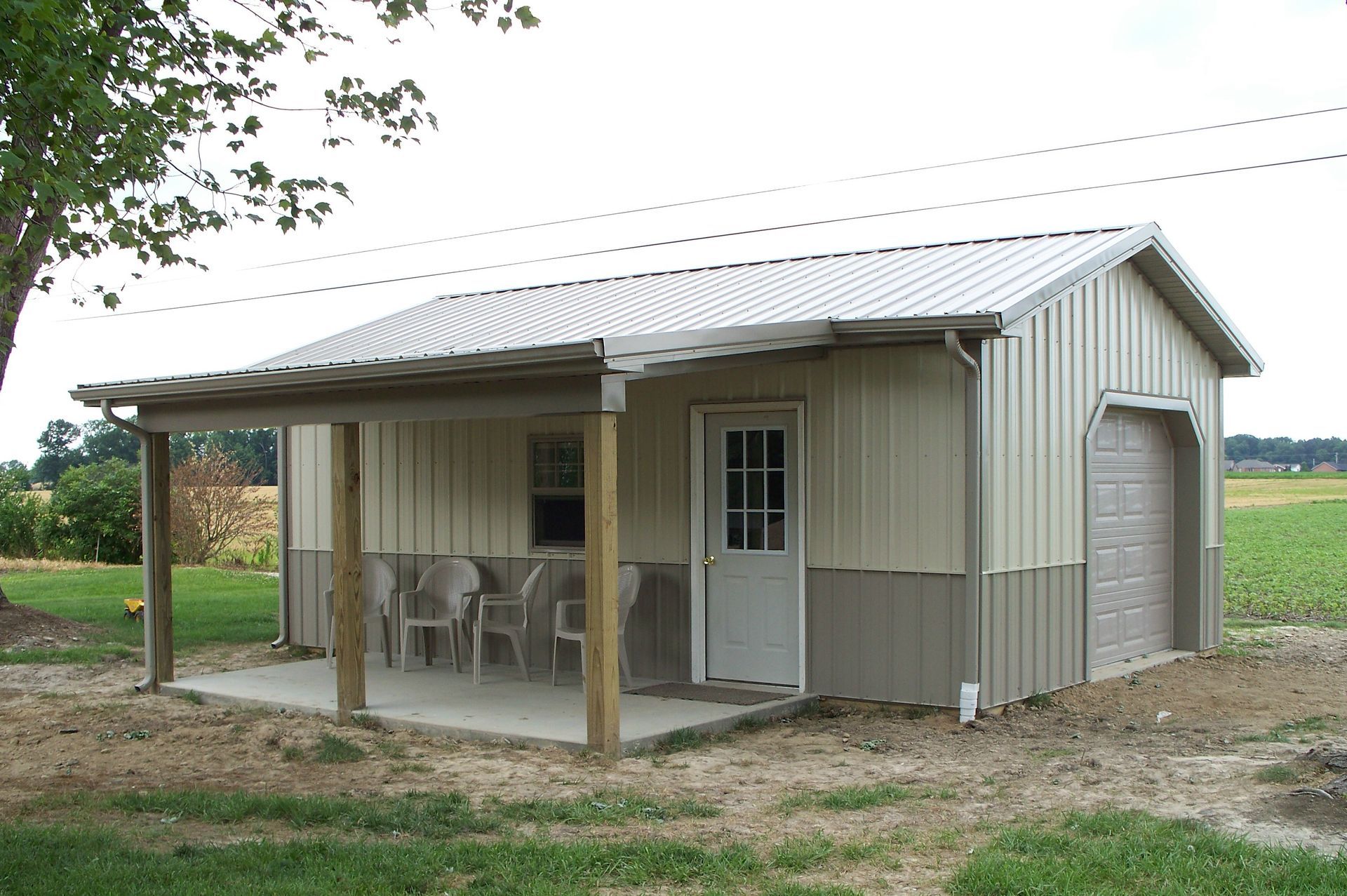 Small beige metal shed with porch, white door, and garage-style side door in a grassy rural yard