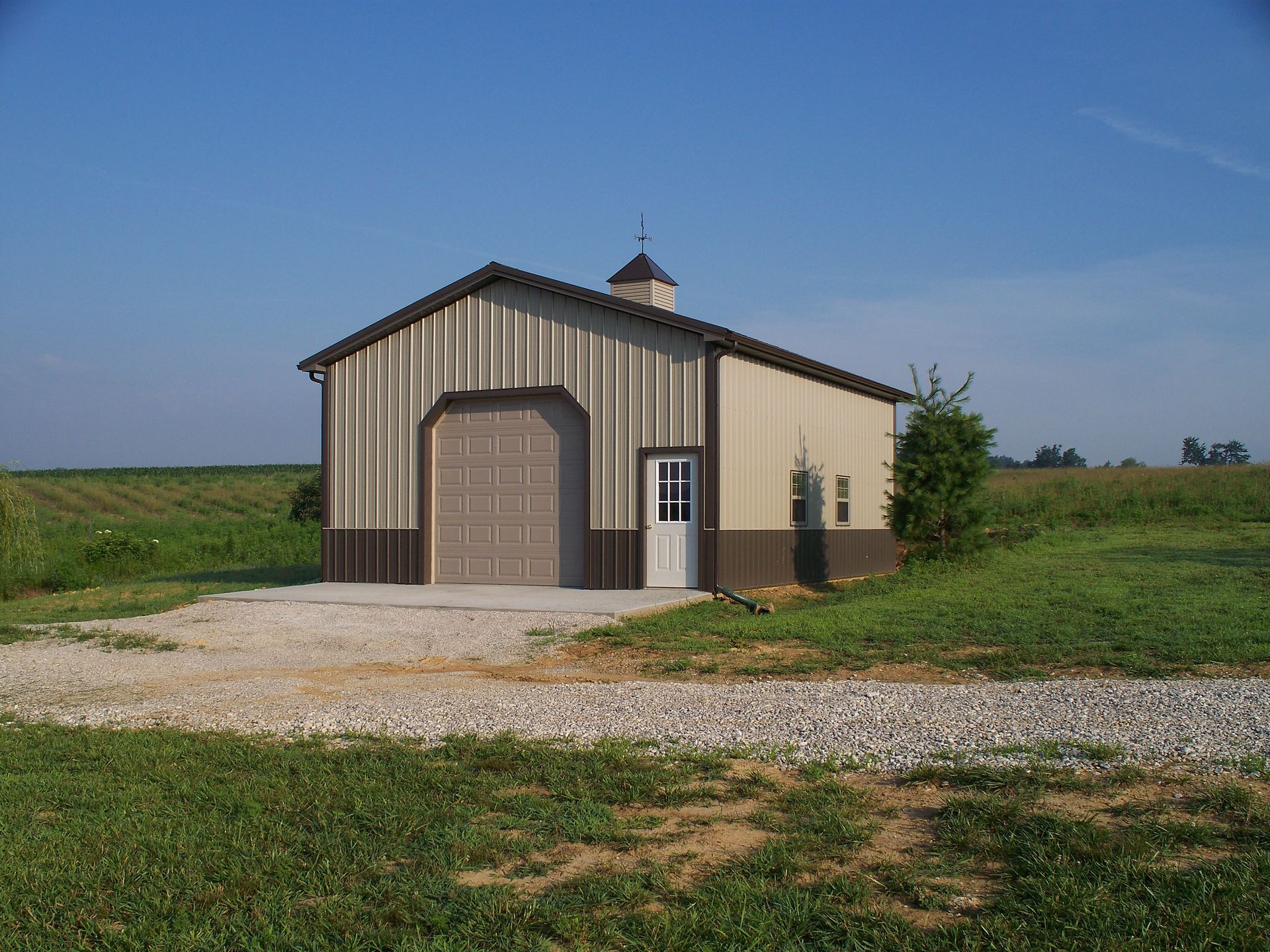 Small beige metal barn with a garage door, set on a gravel drive in a rural field under a clear blue sky.