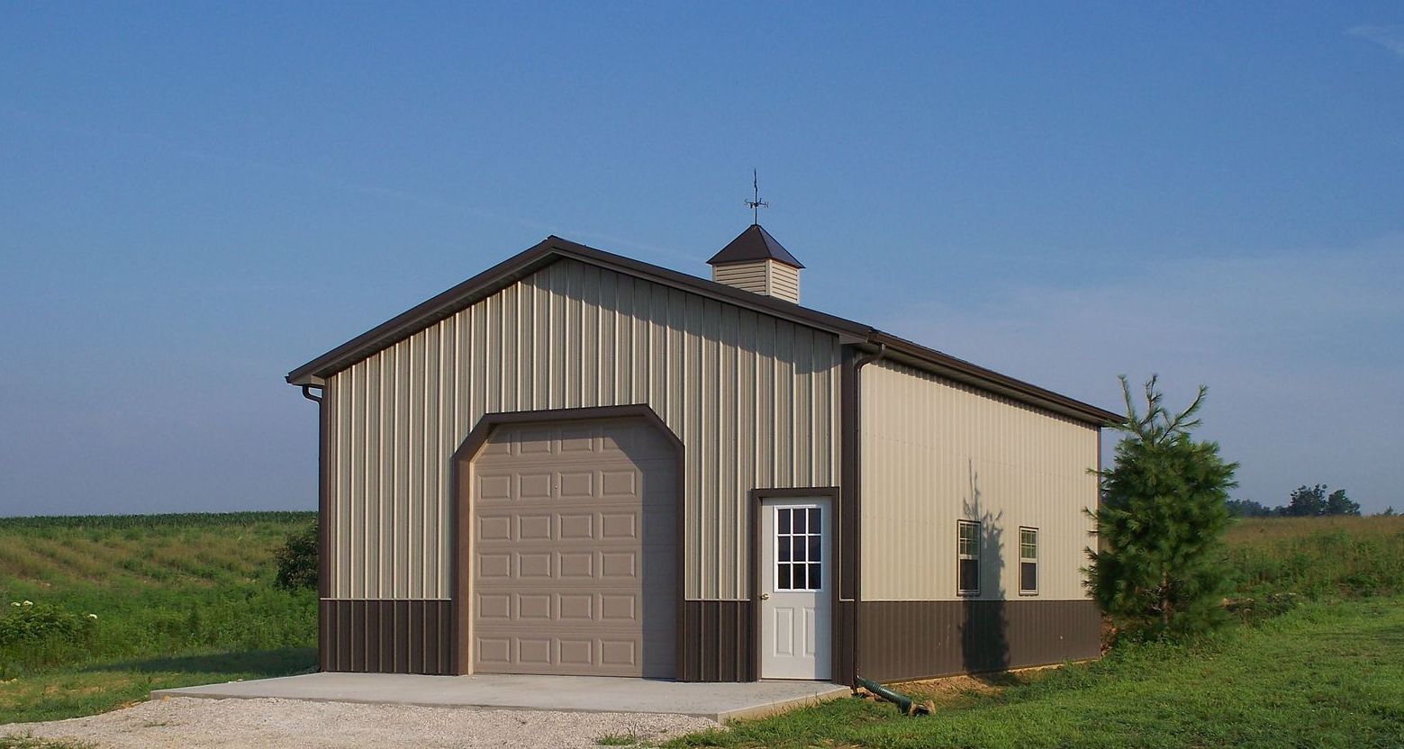 Small beige barn with brown trim and a large garage door, set in a grassy field under a blue sky