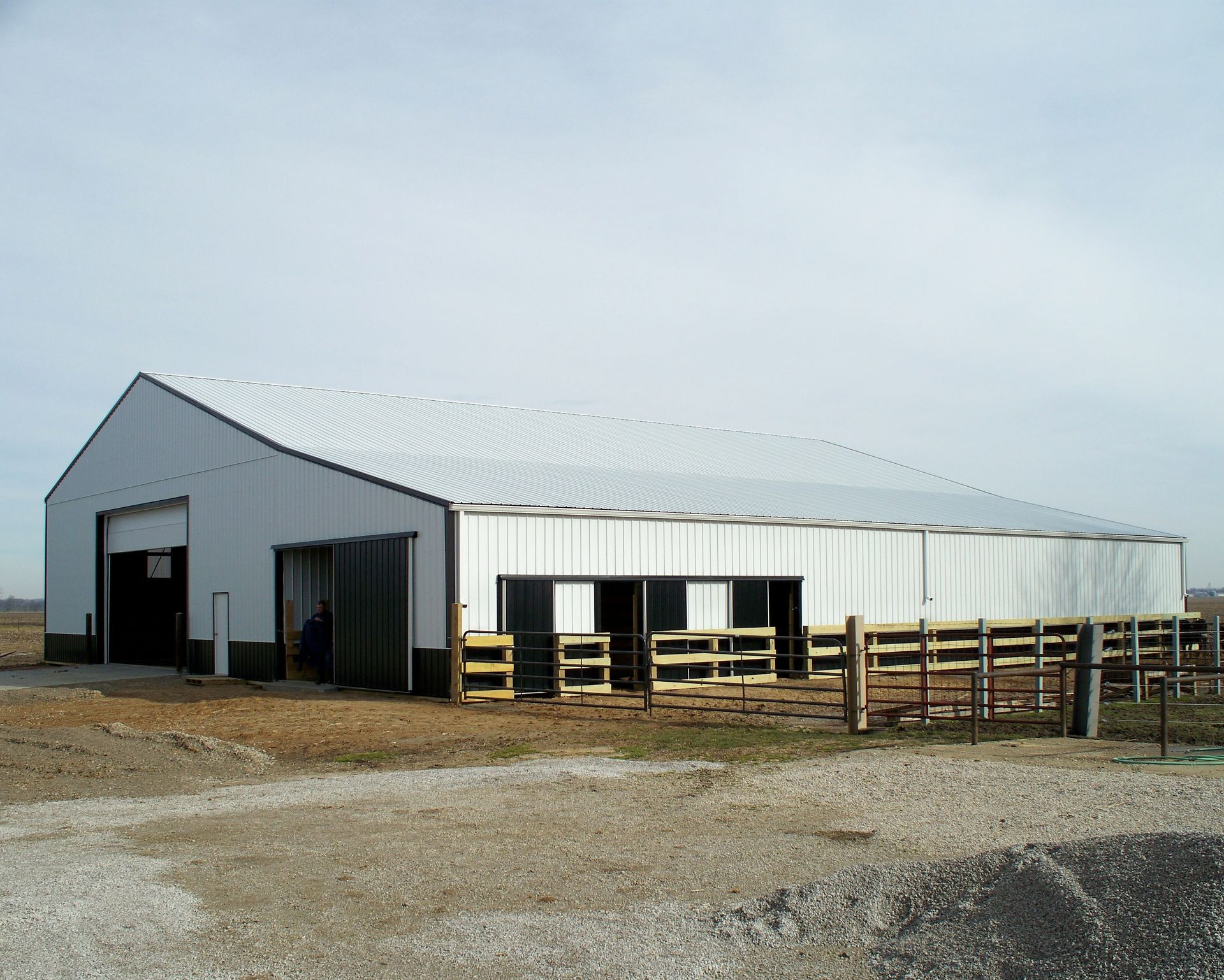 White metal barn with open front and fenced pens on a gravel lot under a cloudy sky