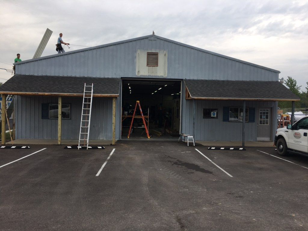 Gray warehouse under repair with ladders and a worker on the roof, seen from a parking lot