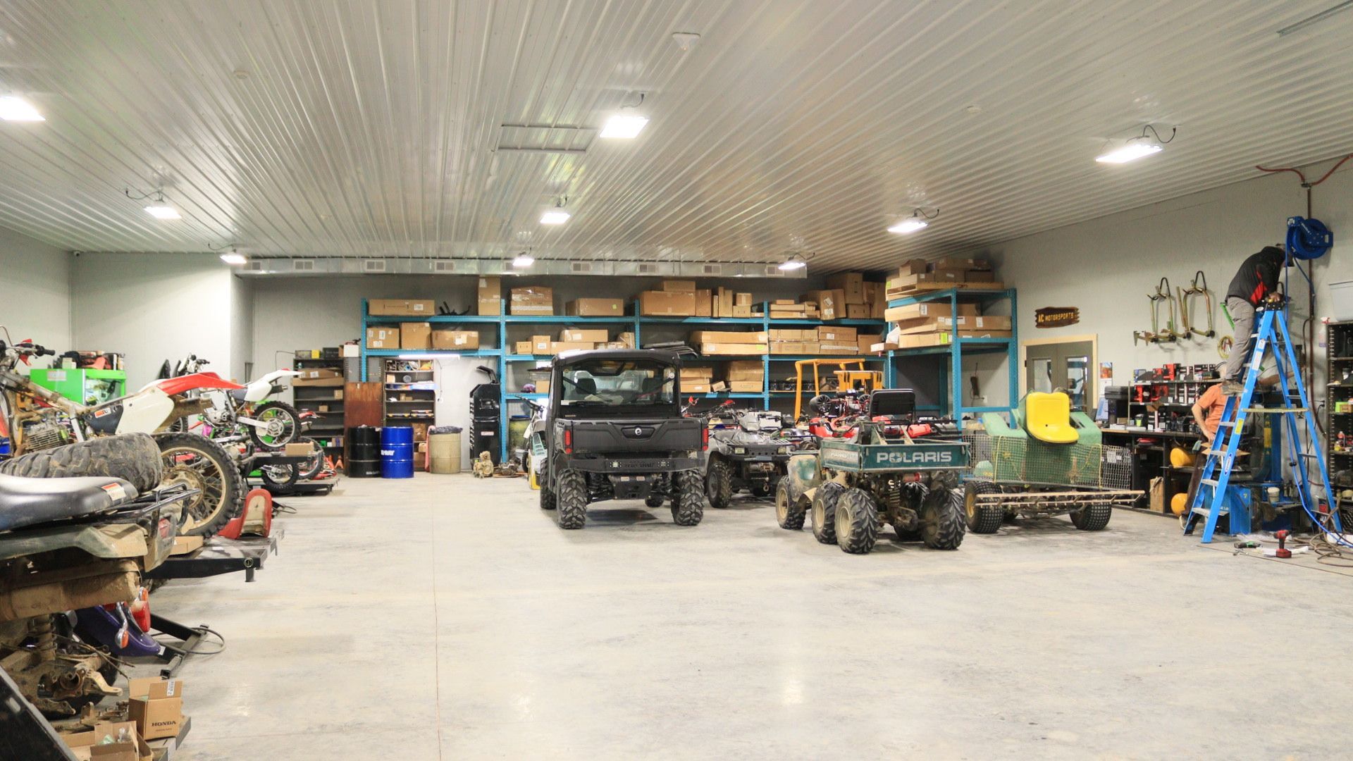 Wide garage workshop with several ATVs, tools, shelves, and a blue ladder under bright ceiling lights