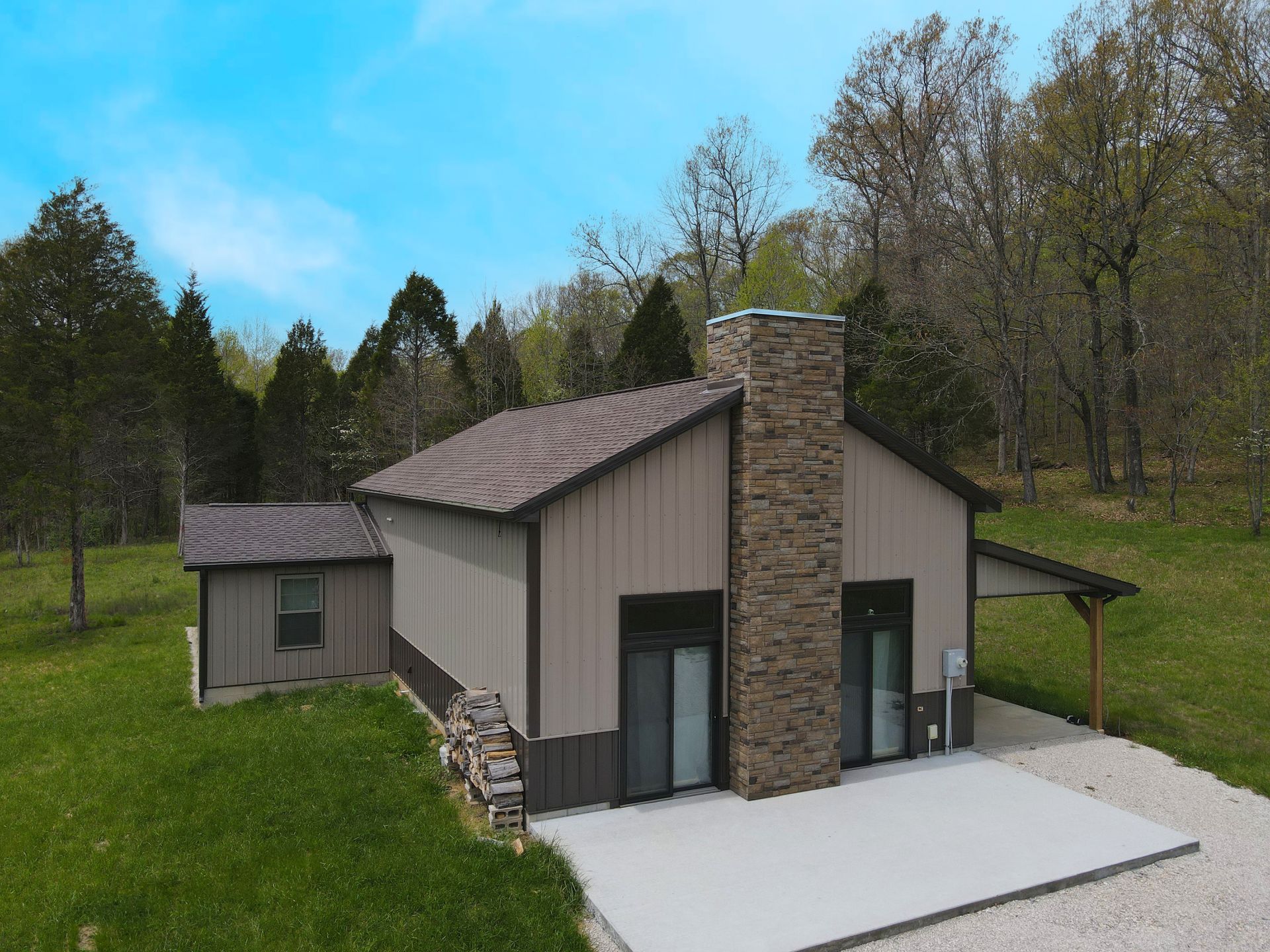 Small modern house with stone chimney, gray siding, and a concrete patio on a grassy hillside.