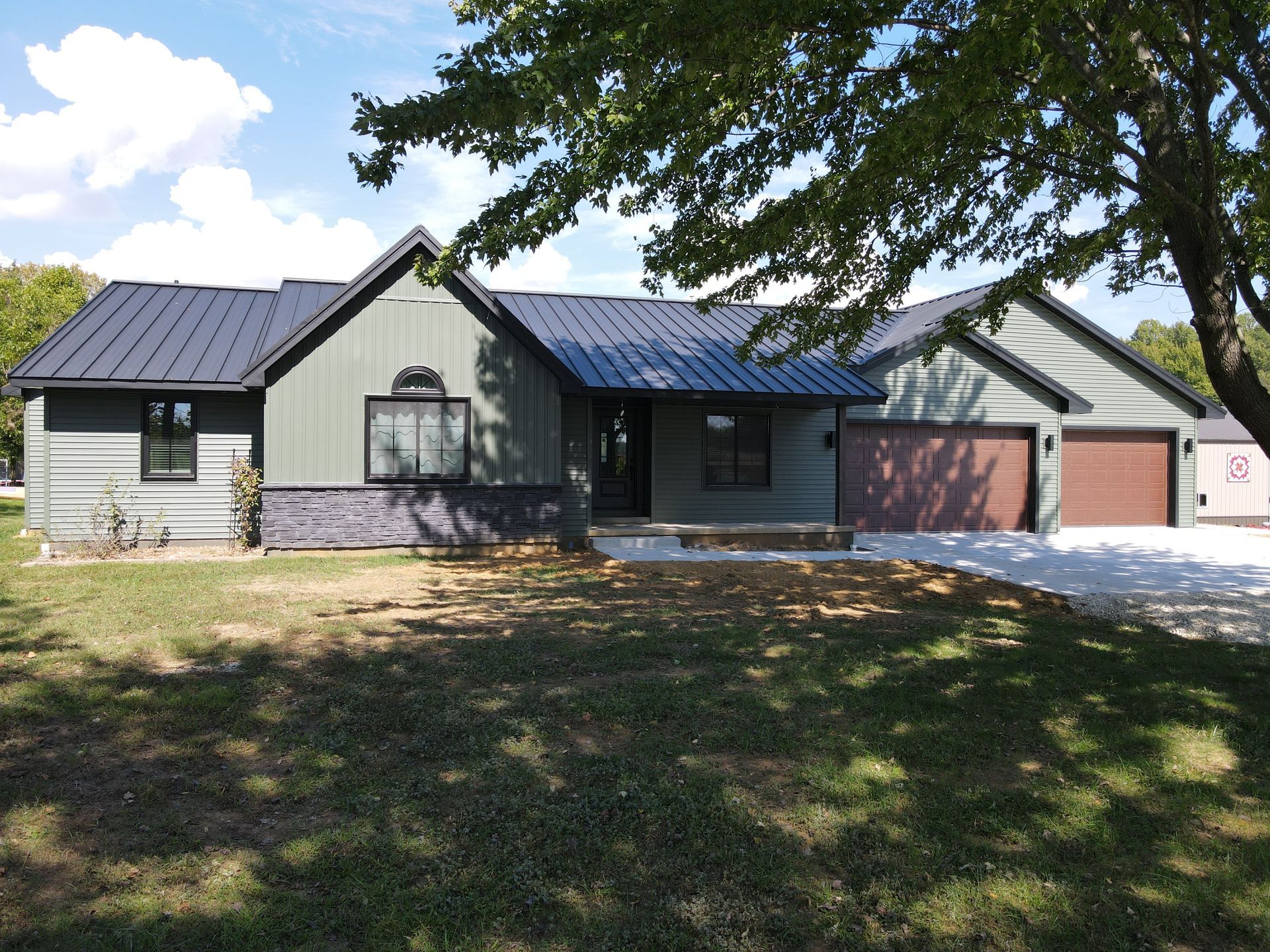 Single-story house with gray siding, dark roof, and attached garage under a tree in a sunny yard