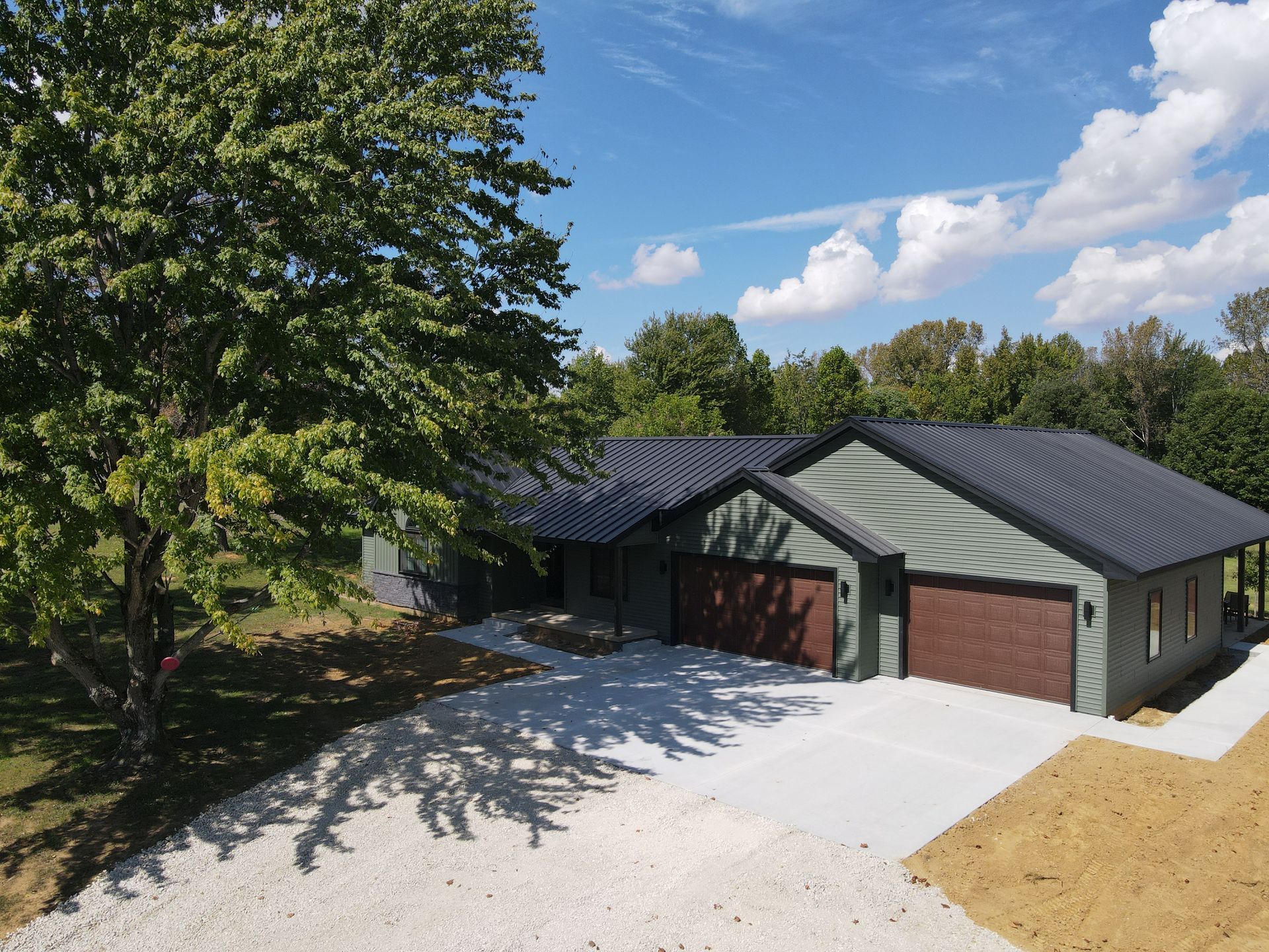 Suburban house with three garage doors, gray siding, and a large tree casting shadows on the driveway.