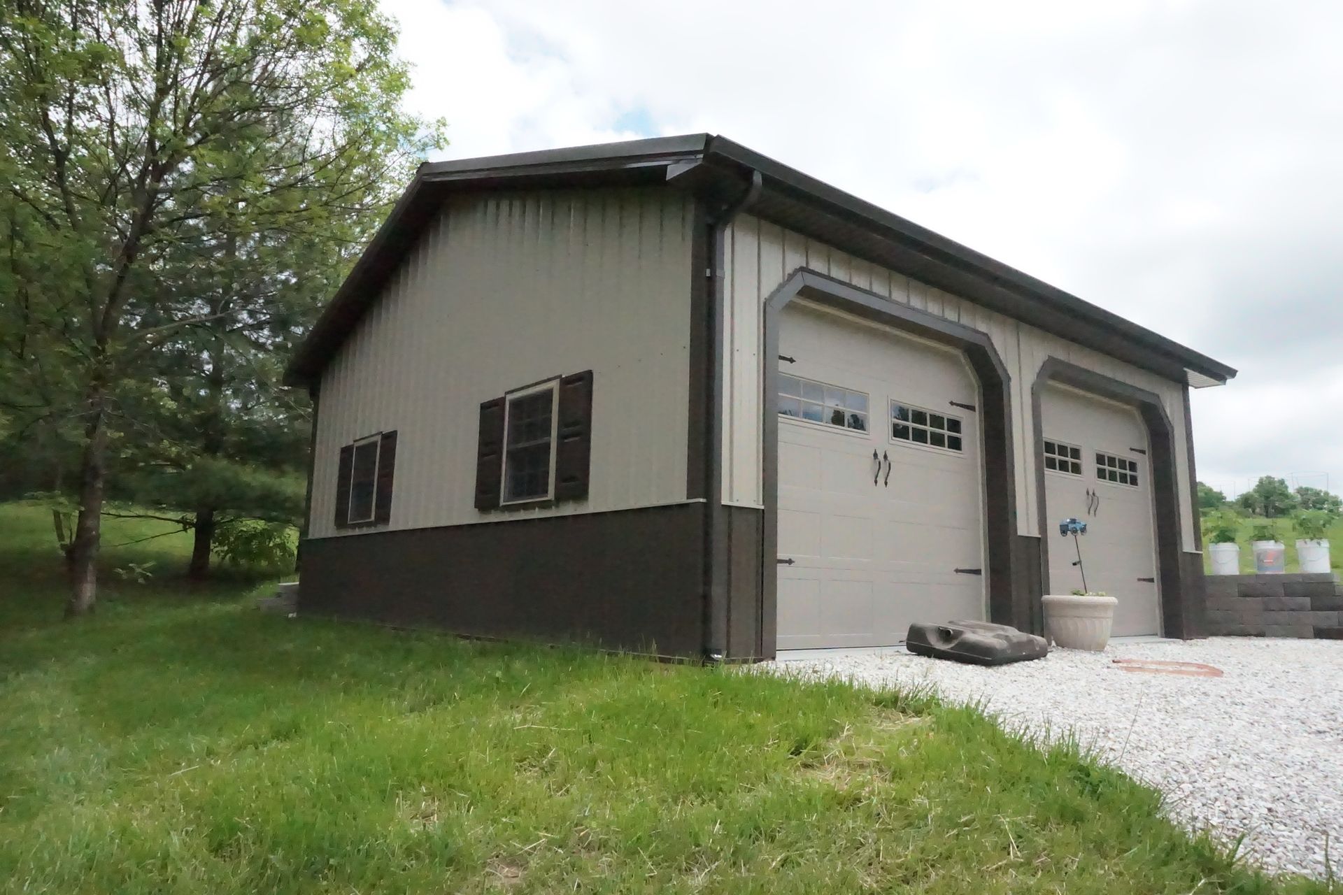 Detached beige garage with two white doors, black trim, and gravel driveway beside a grassy yard