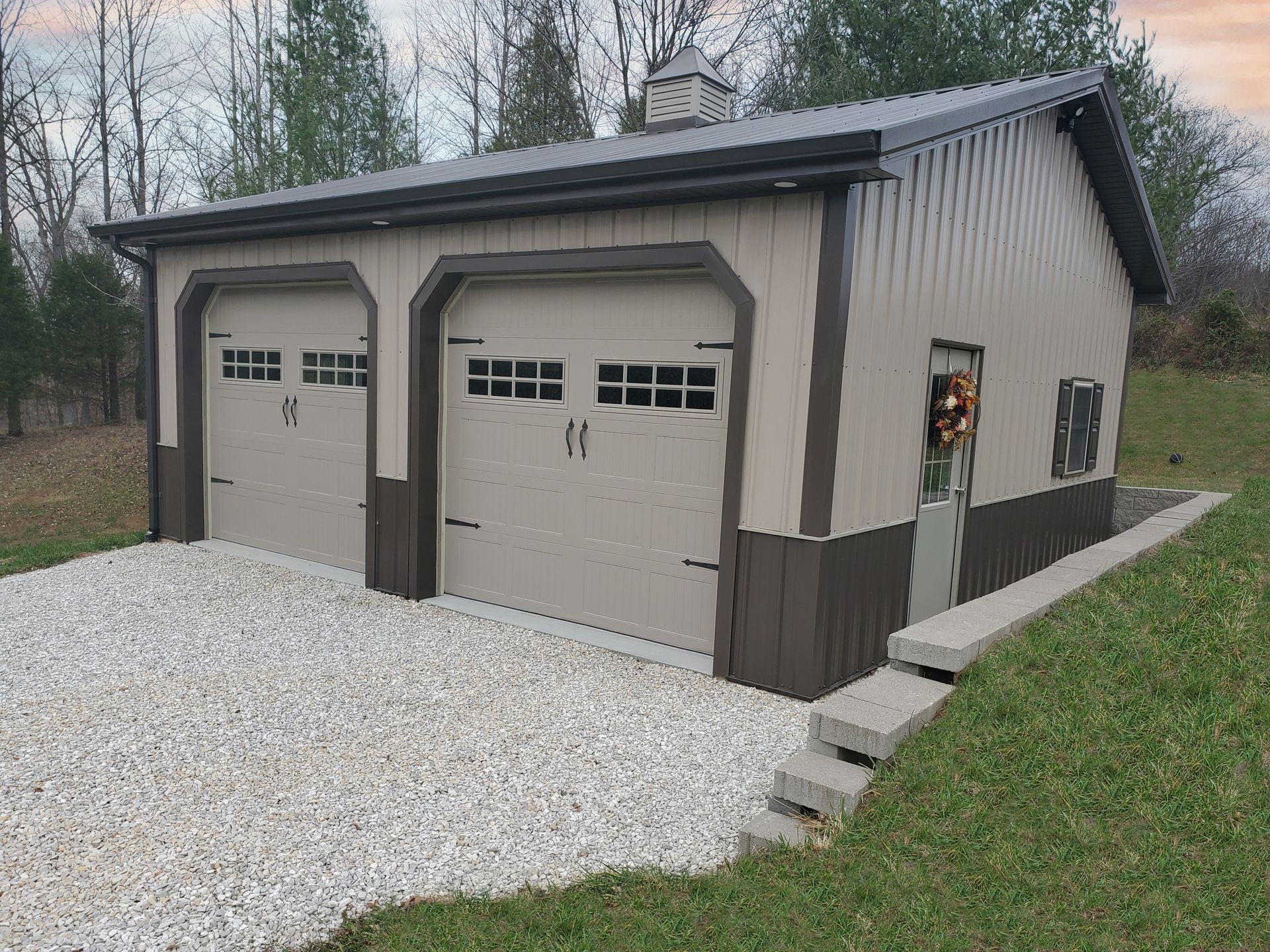 Detached two-car garage with tan doors, brown trim, and gravel driveway beside a grassy slope