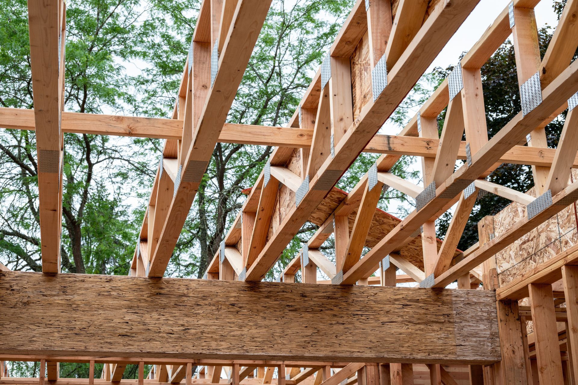 Wooden floor and roof framing under construction, with exposed timber joists and trees visible. Wooden floor and roof framing under construction, with exposed timber joists and trees visible.
