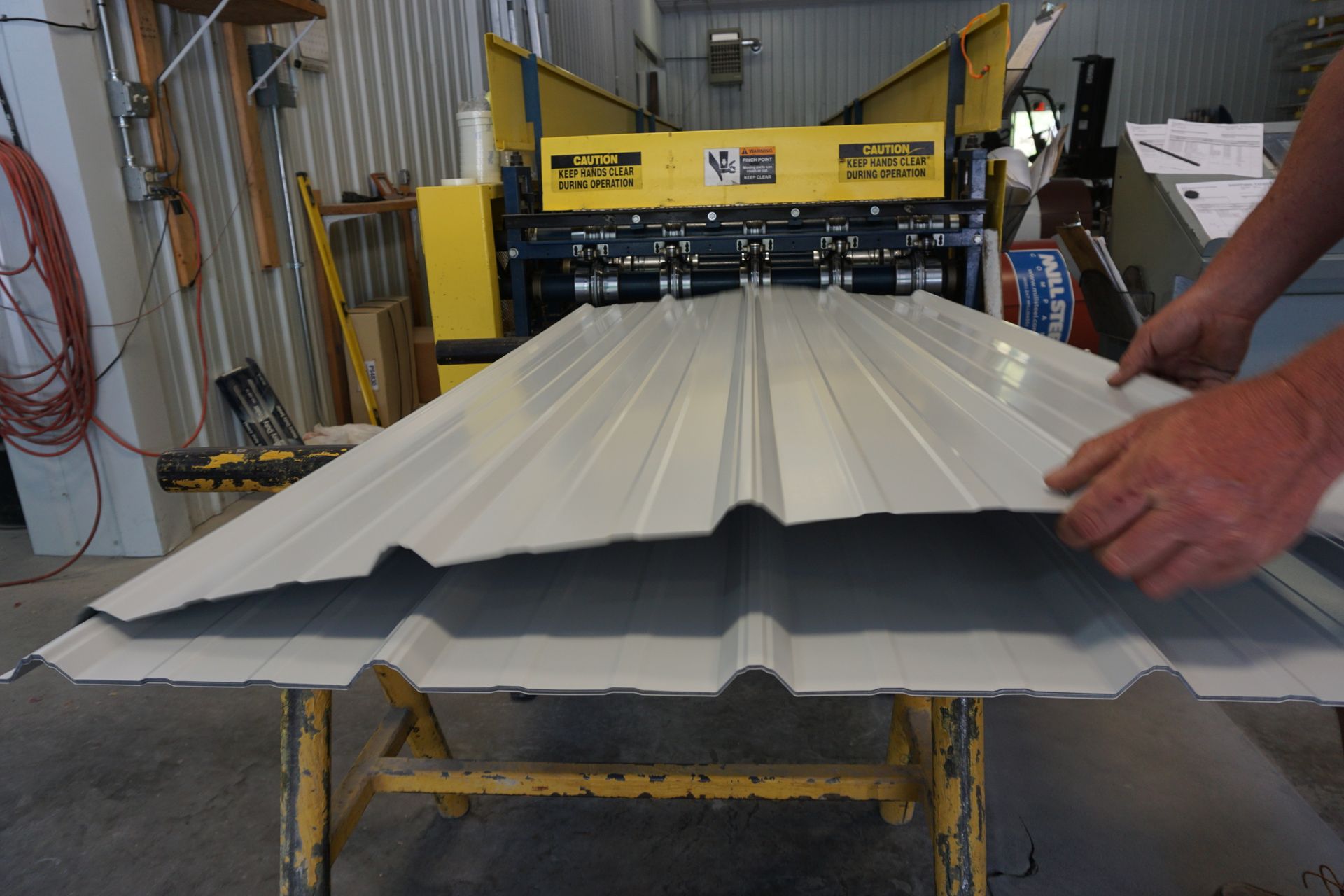 Worker handling a white corrugated metal sheet at a roll-forming machine in a workshop