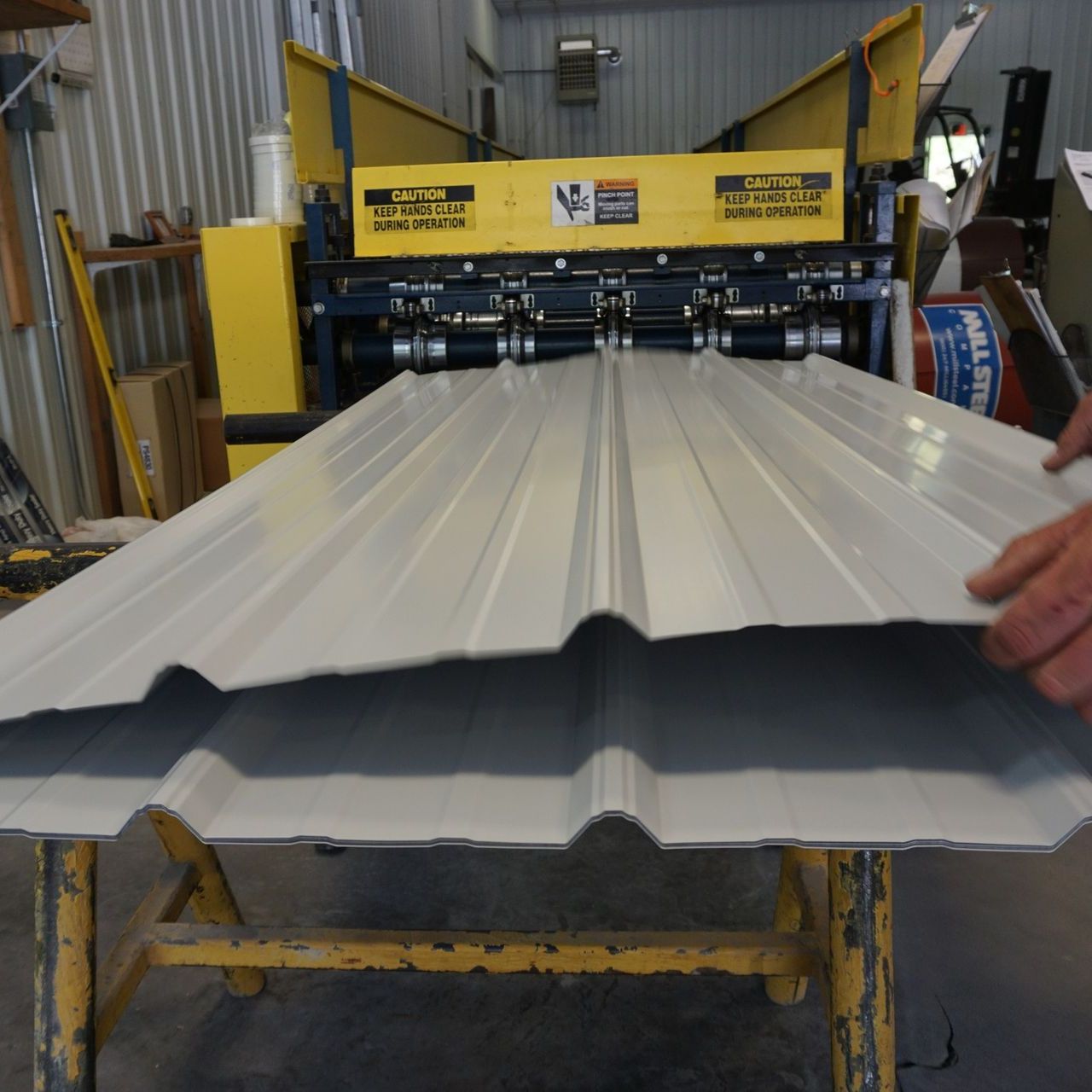 White corrugated metal roofing panel being fed through a yellow roll-forming machine in a workshop