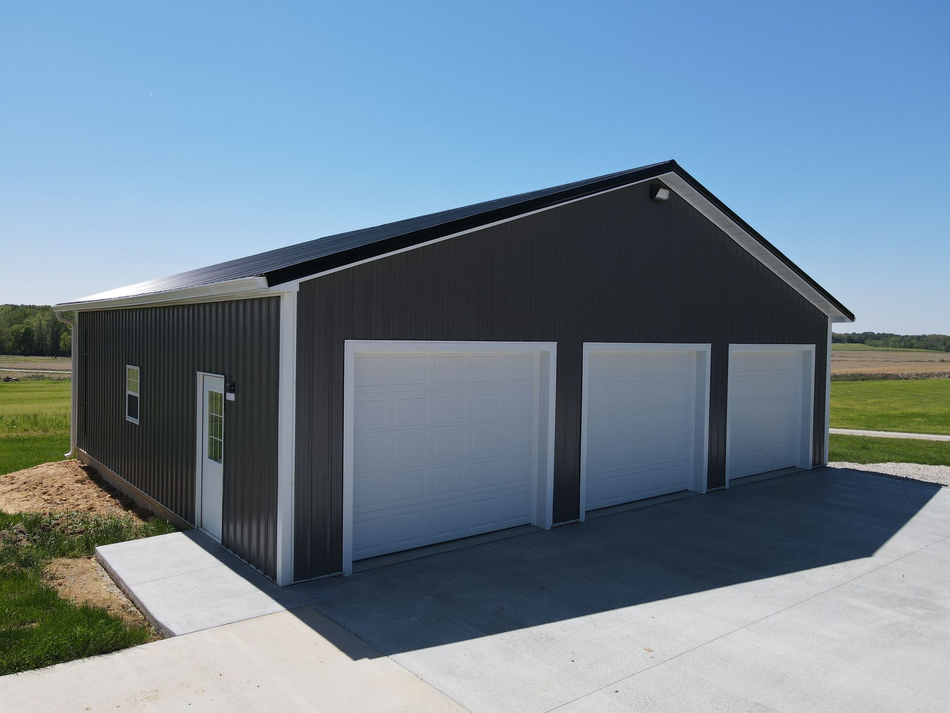 Gray metal garage building with three white roll-up doors on a rural lot under a clear sky