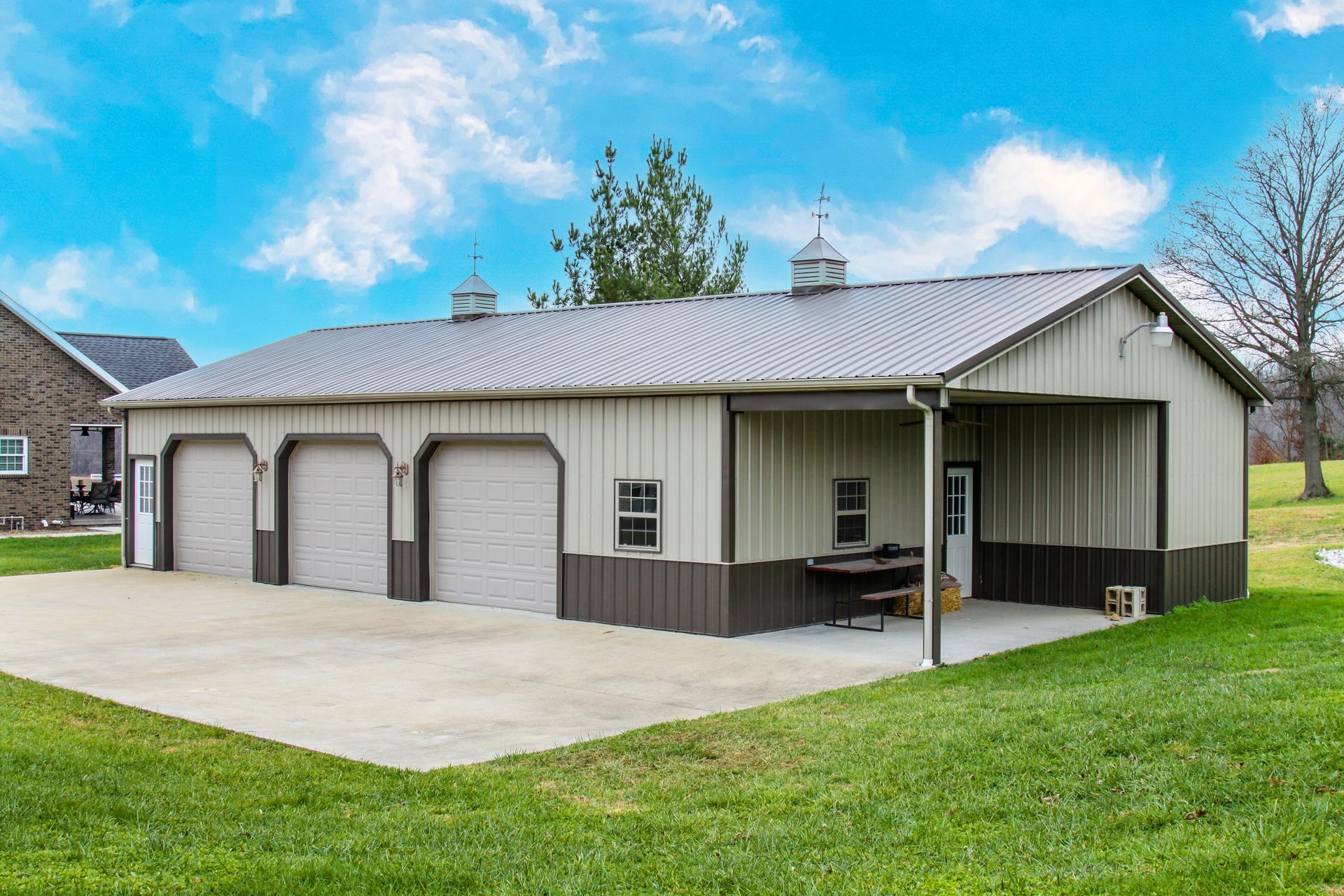 Detached metal garage with three white doors, covered side porch, and paved driveway beside a grassy yard