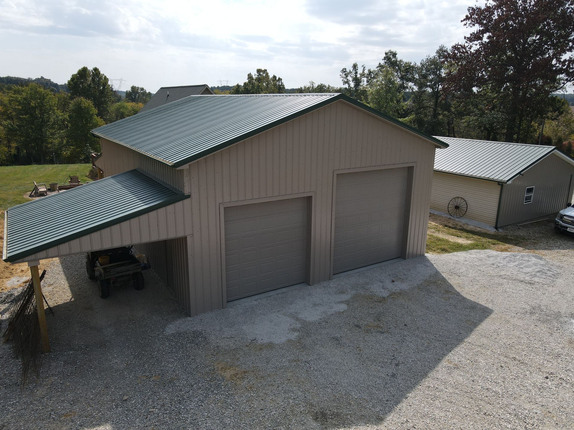 Gray metal barn with two garage doors and a carport on a gravel driveway, surrounded by trees.