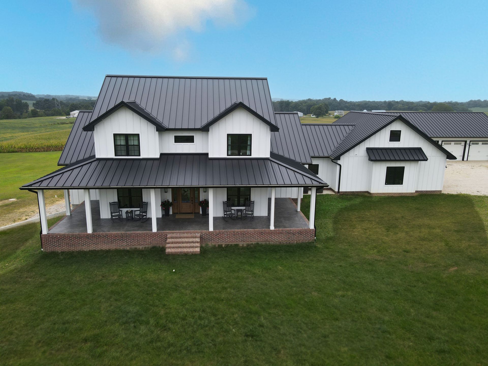 White farmhouse with metal roof and covered porch beside a detached garage on a grassy rural lot.