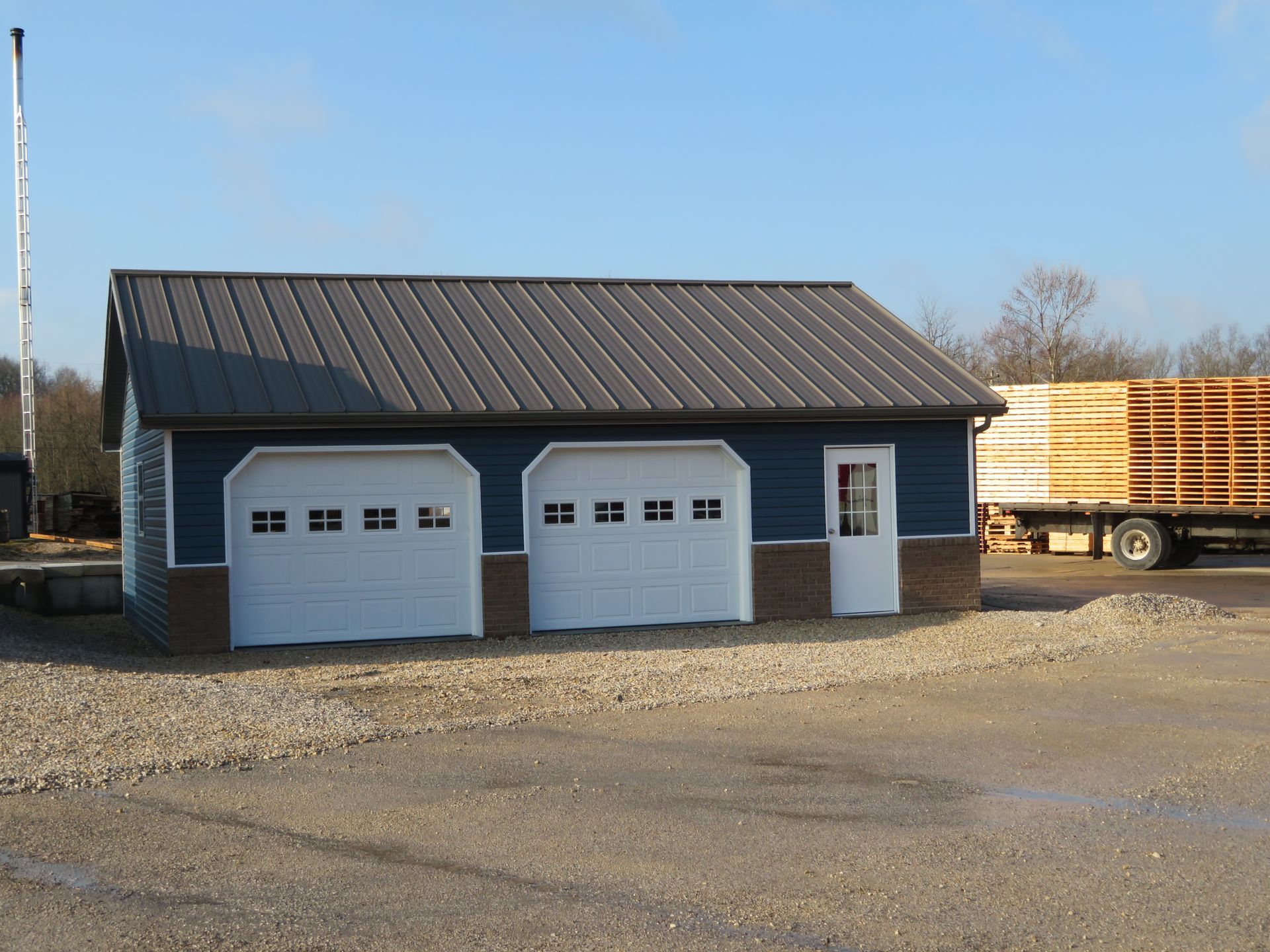 Blue garage with two white doors beside a parked trailer on a gravel lot under a clear sky
