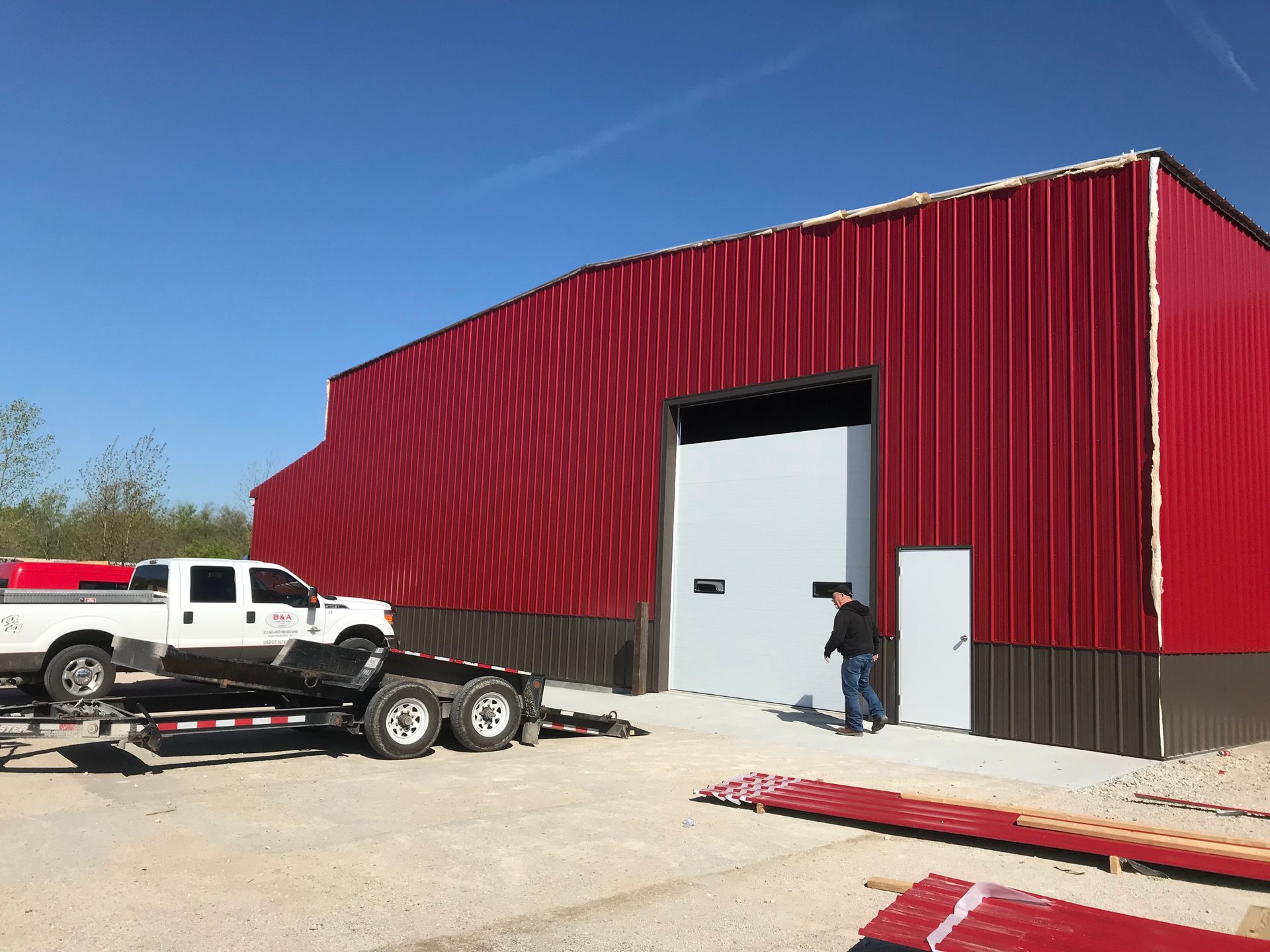 Red metal warehouse with a white roll-up door; a worker stands at the entrance beside a trailer and truck.