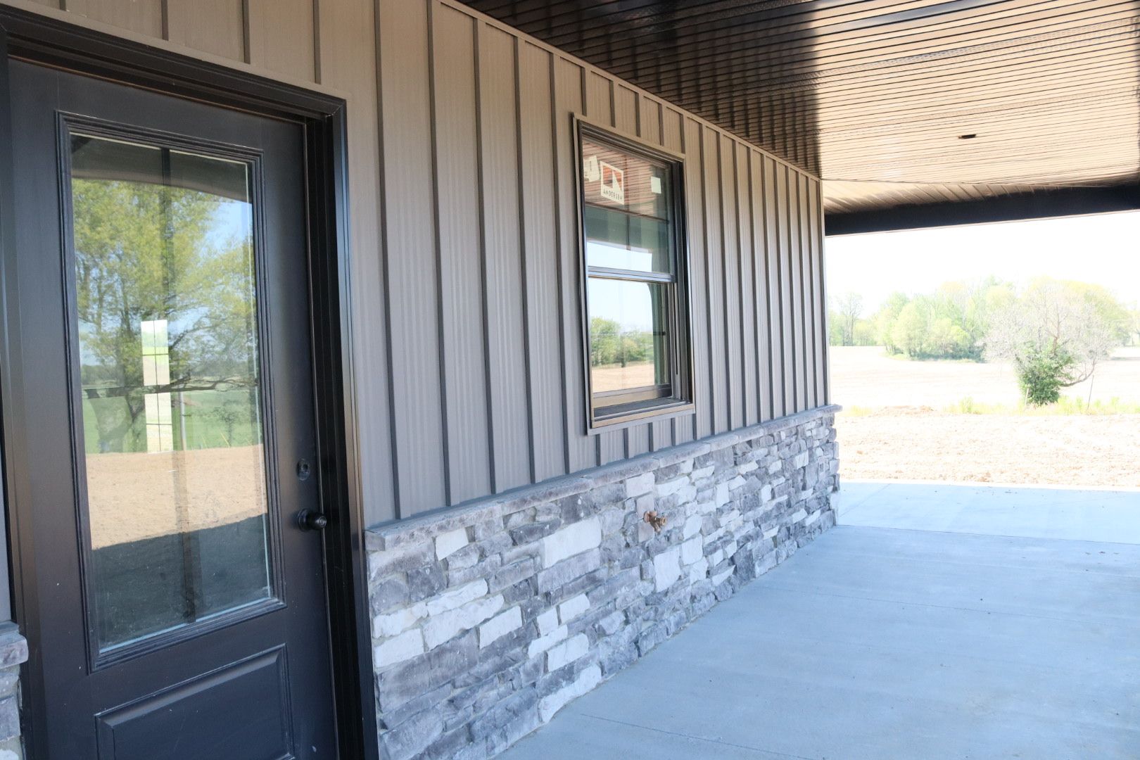 Covered porch with stone and vertical wood siding, a black door, and windows overlooking an open field