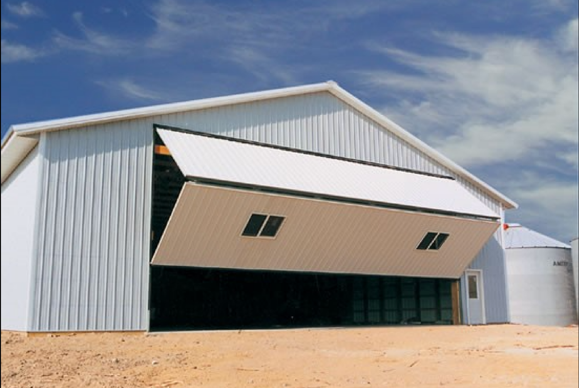 Large white metal hangar with a partially open front door in a sandy industrial area