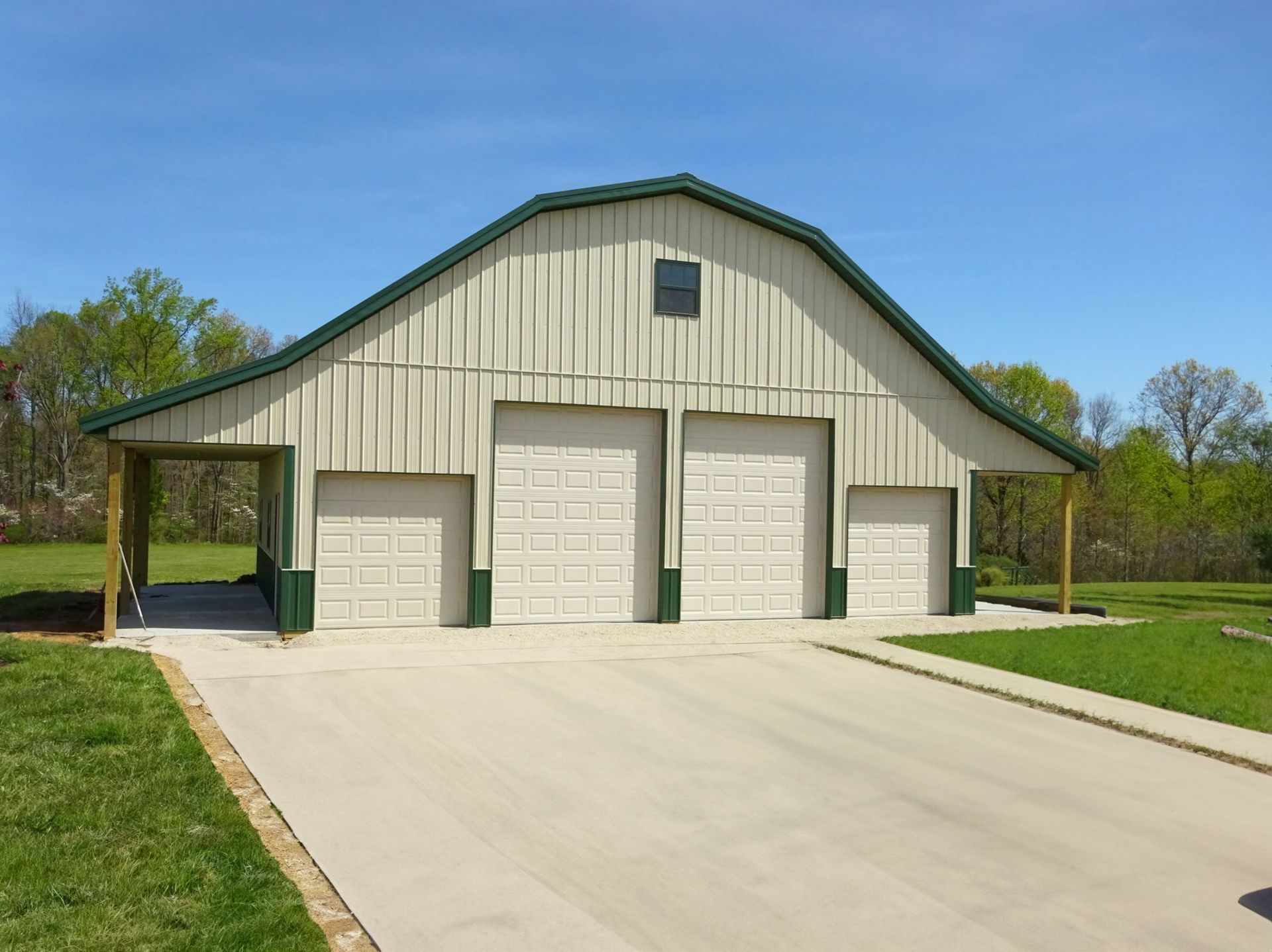 Large beige barn with green trim and garage doors beside a concrete driveway on a sunny day