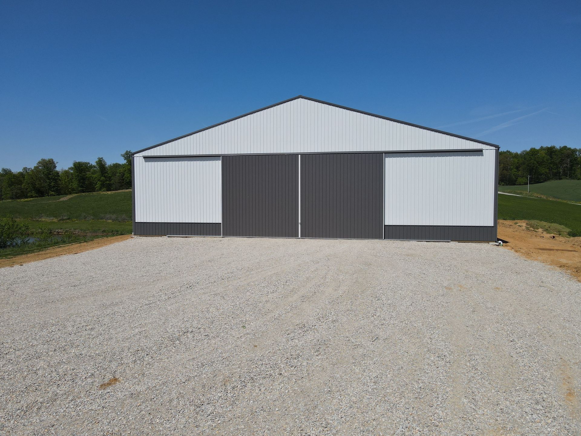 Large gray metal barn with white sliding doors on a gravel lot under a clear blue sky