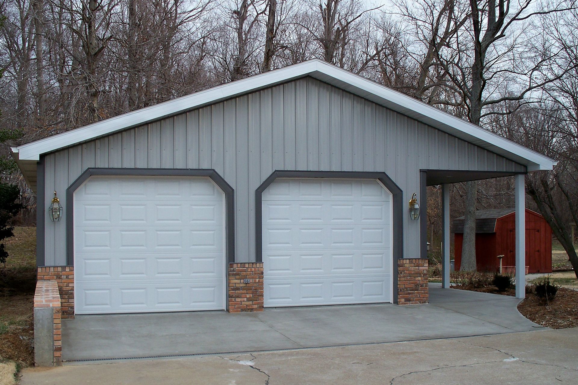 Two-car garage with light gray siding, white doors, and a small side porch in a wooded yard.