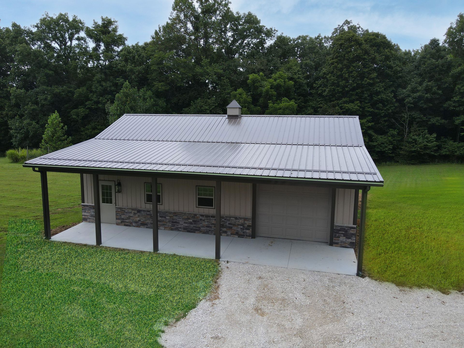 Small gray house with a metal roof, front porch, and gravel driveway beside a grassy yard and trees
