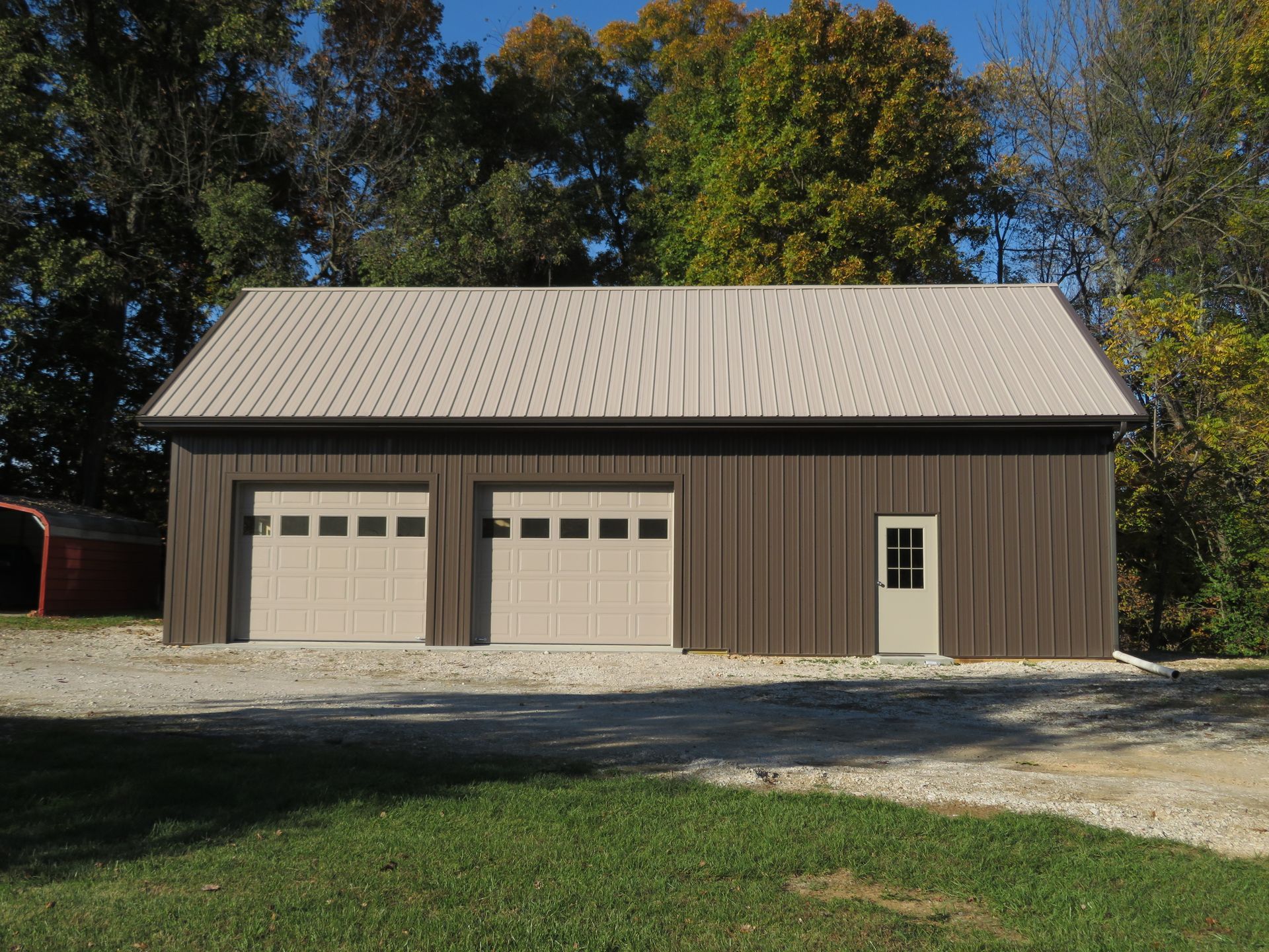 Brown garage with two beige doors and a side entry door, set against trees and a gravel driveway.