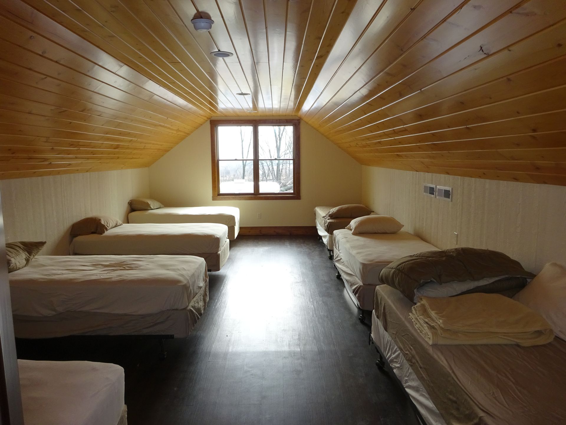 Dorm-style bedroom with four single beds under a sloped wood-paneled ceiling and a central window.