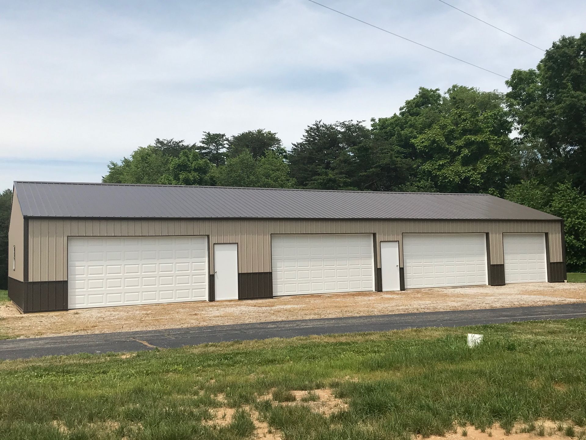 Long beige garage building with four white doors and dark roof beside a grassy roadside