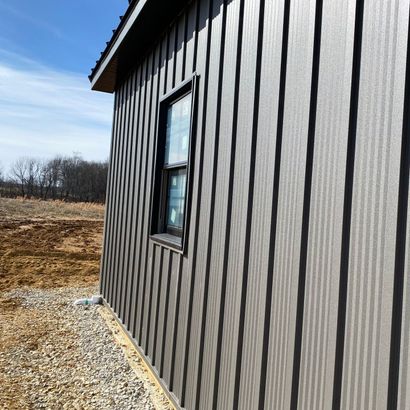 Side of a gray metal building with a window beside a gravel path in a rural field