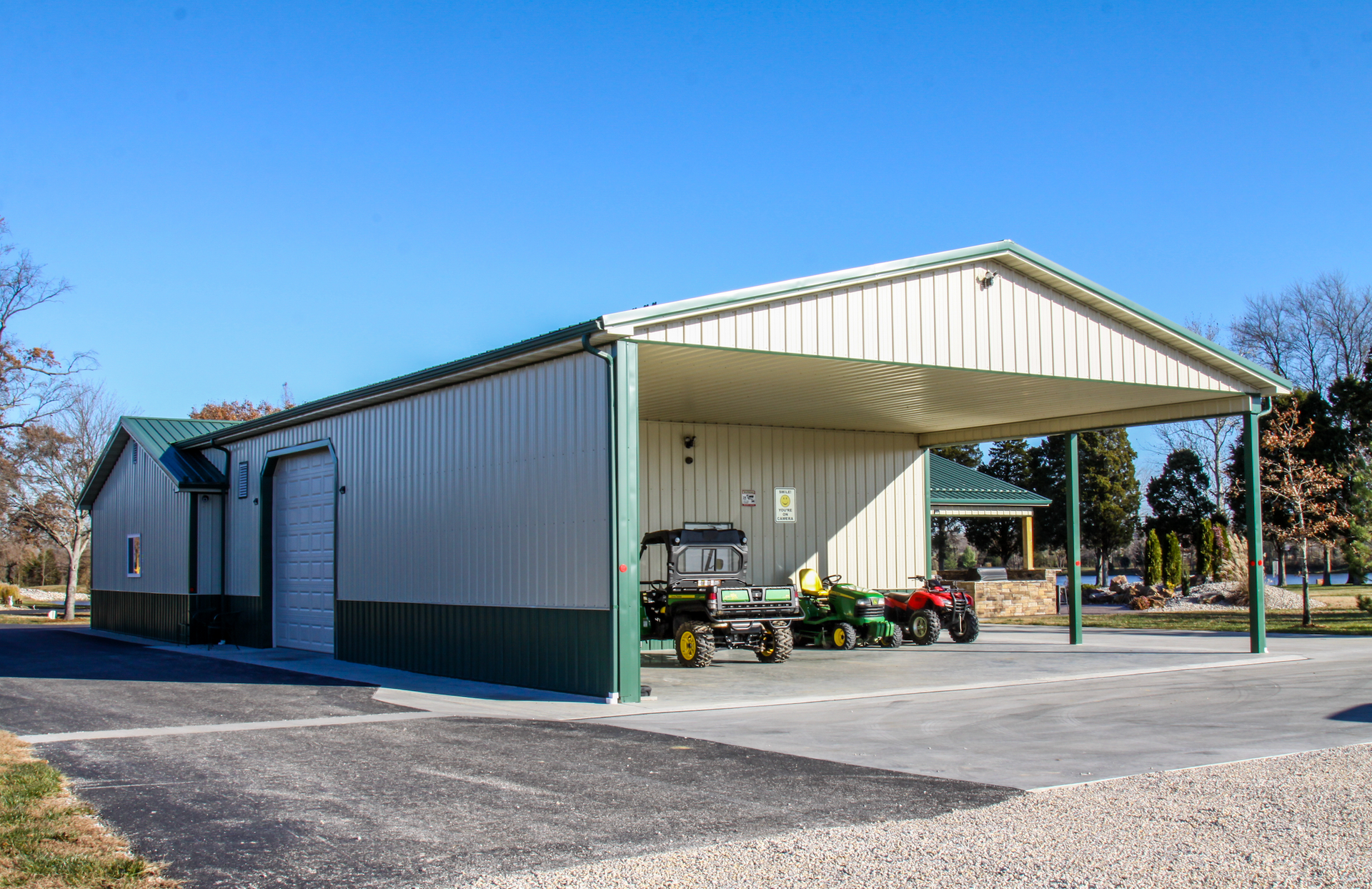 Metal farm shed with open bay and tractors parked underneath on a gravel lot.