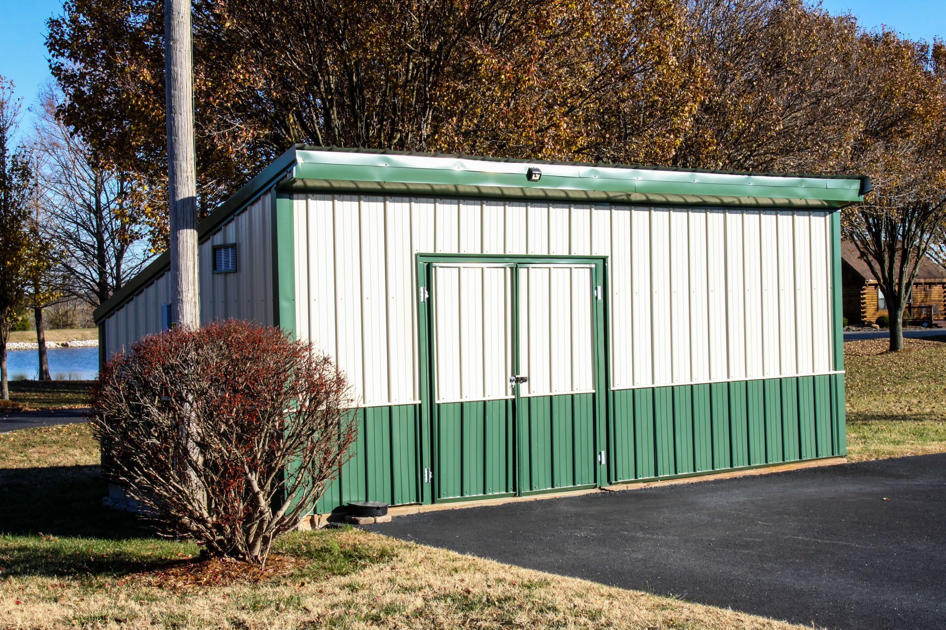 Small green-and-white utility shed beside a parking lot, with trees and water in the background