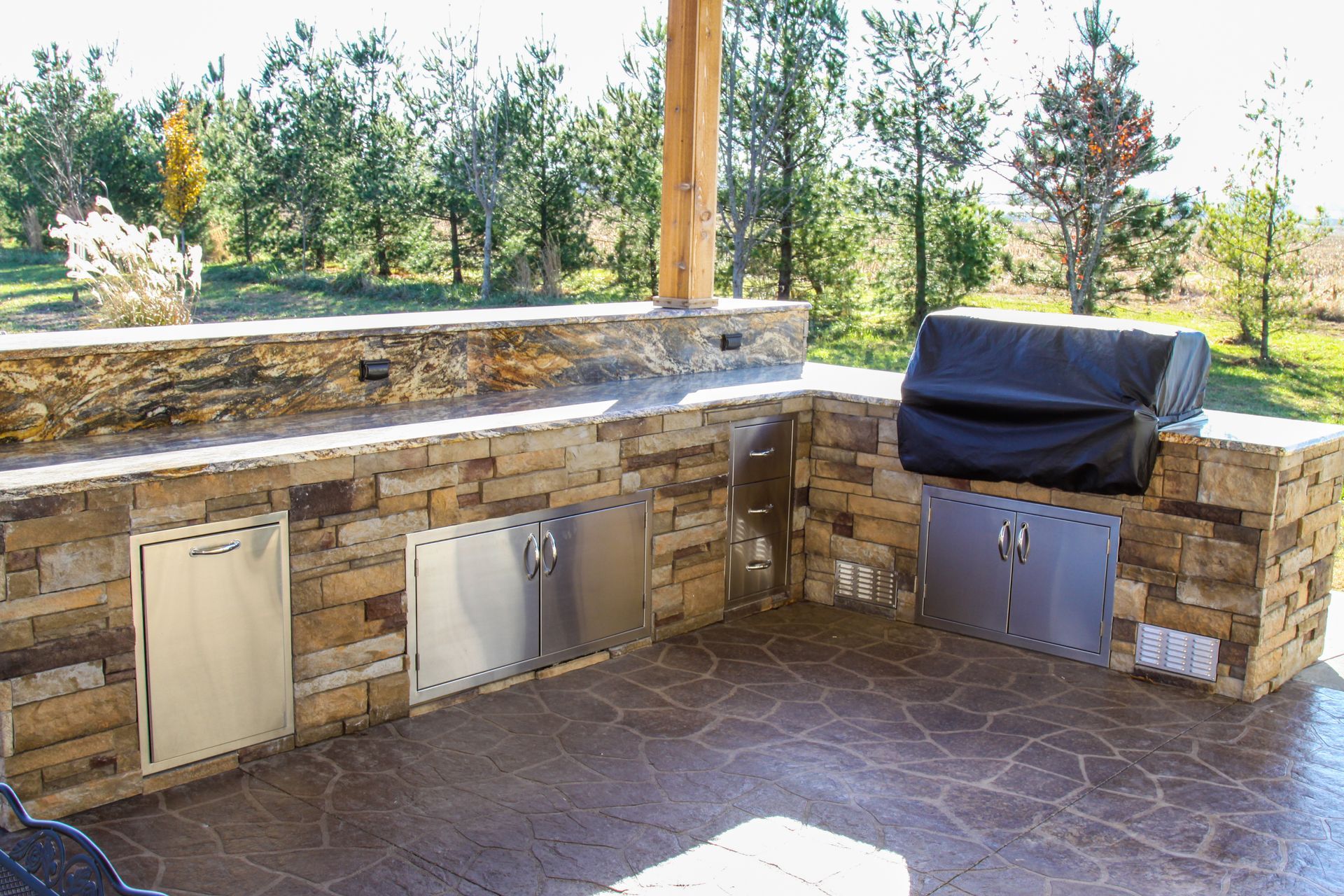 Outdoor stone patio with built-in grill, sink doors, and a covered barbecue in a sunny backyard.