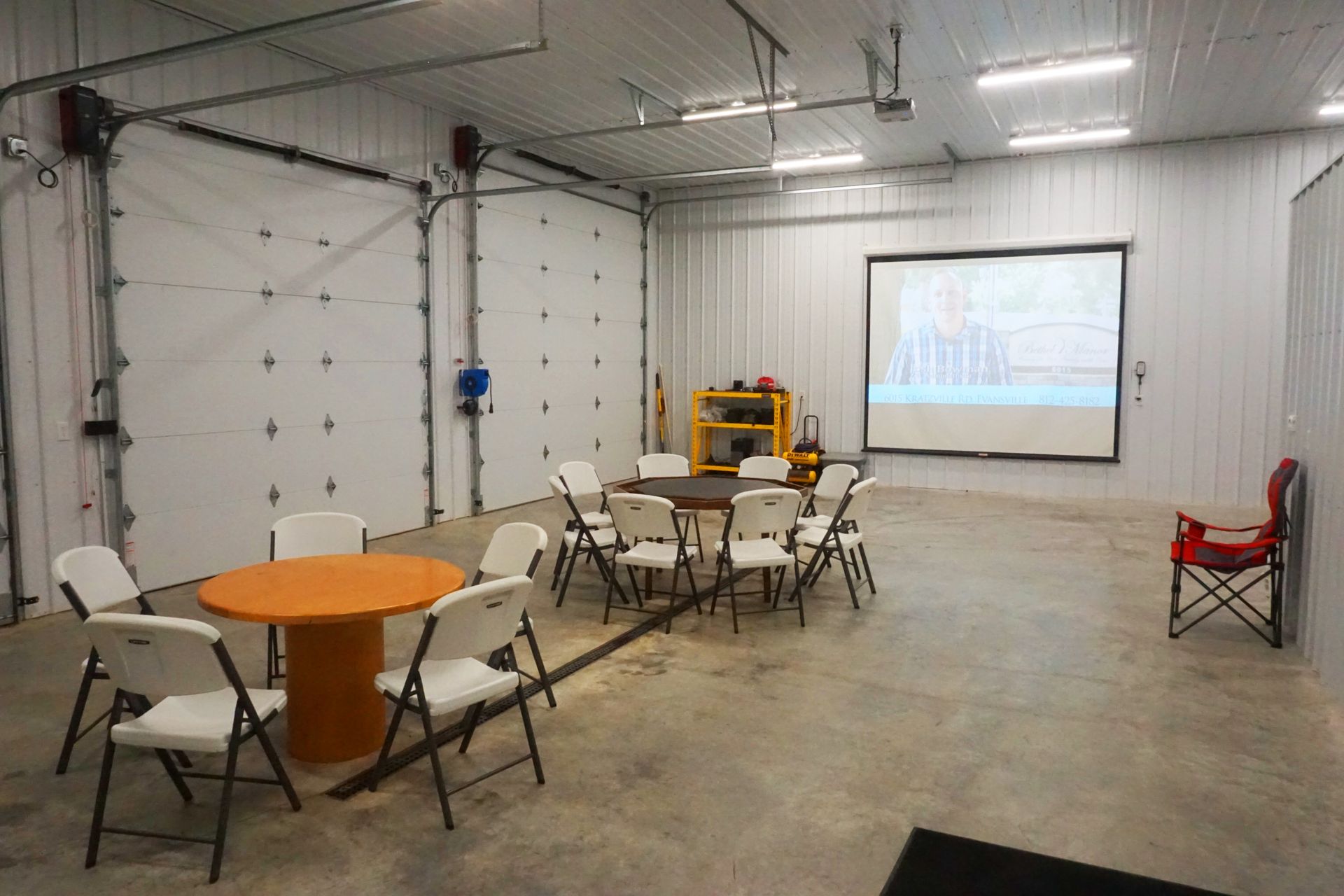 Empty warehouse room with folding tables and chairs, a projector screen, and a small stage setup