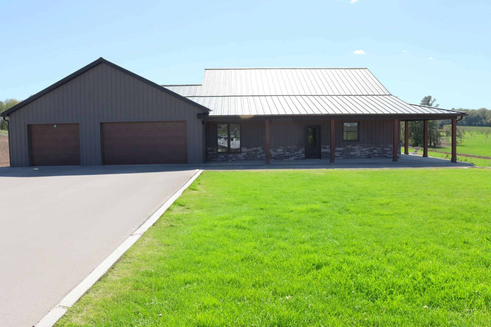 Single-story house with dark siding, white metal roof, driveway, and bright green lawn under a blue sky