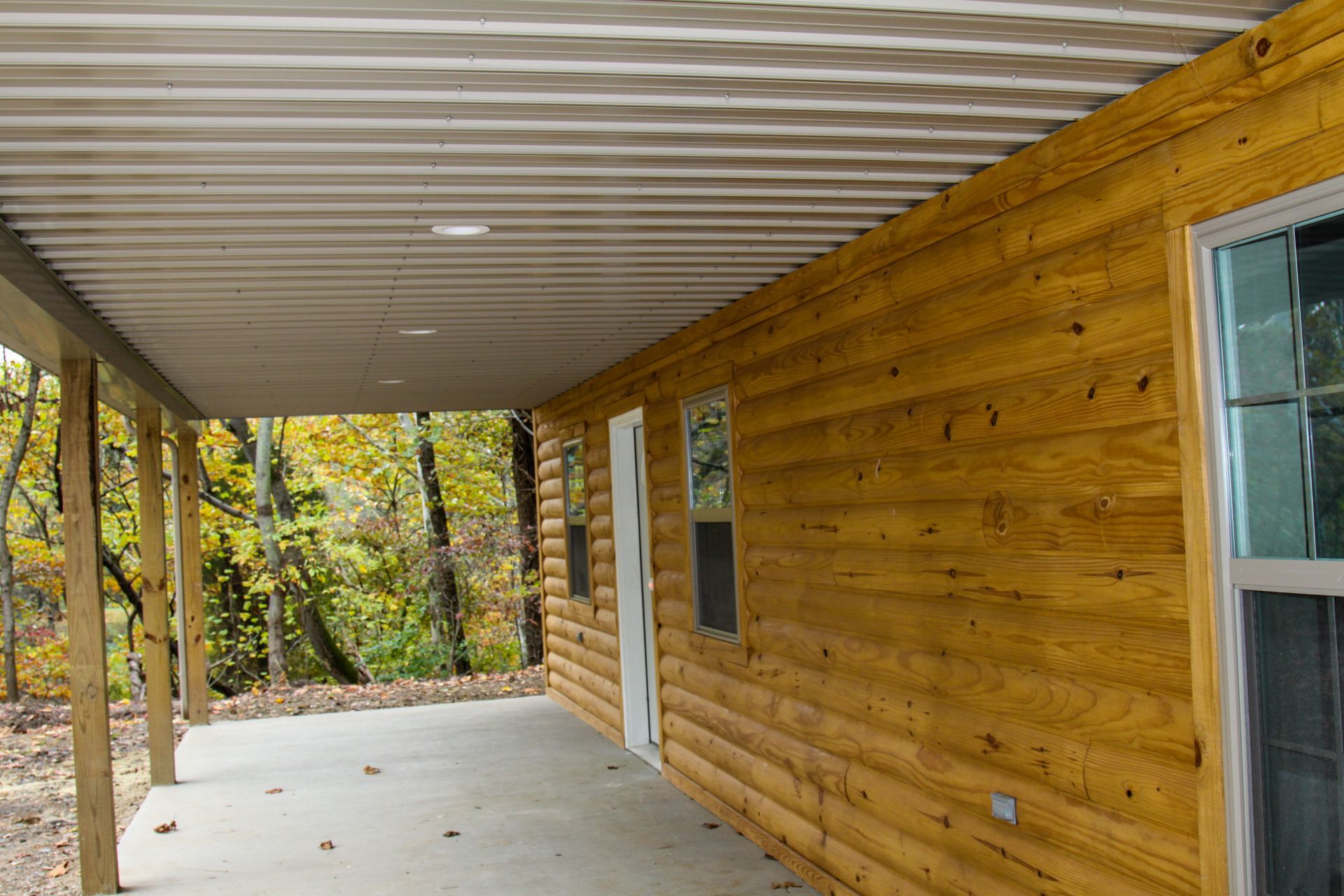 Covered porch beside a log cabin with a concrete floor and forest view beyond the posts