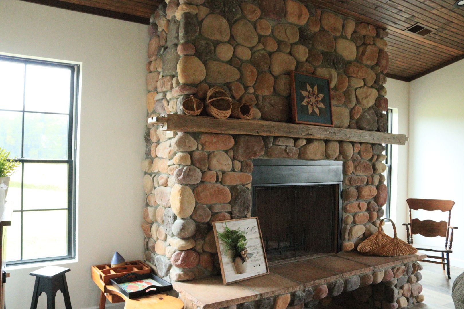 Stone fireplace with a rustic mantel in a bright room, flanked by a window and wooden chair.