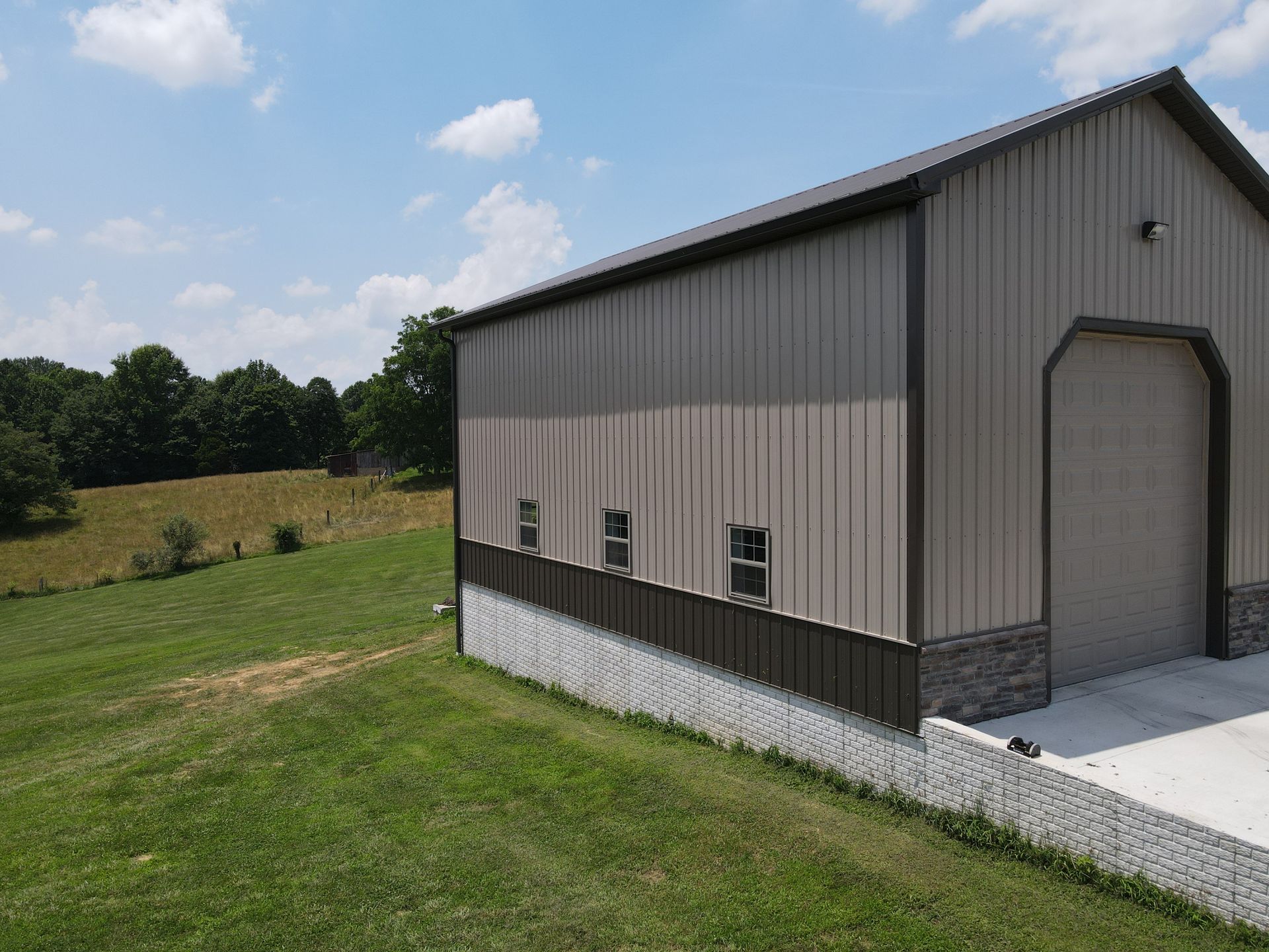 Metal-sided barn with a concrete pad, small windows, and a large roll-up door in a grassy rural field.