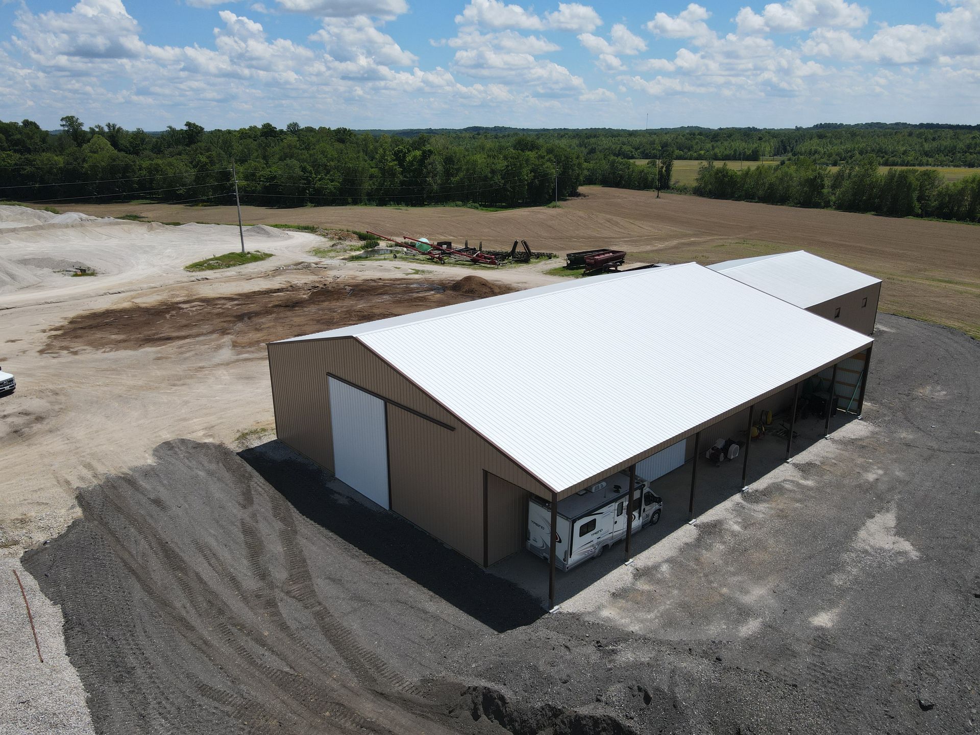 Rural industrial building with a white metal roof beside gravel lots and fields under a partly cloudy sky