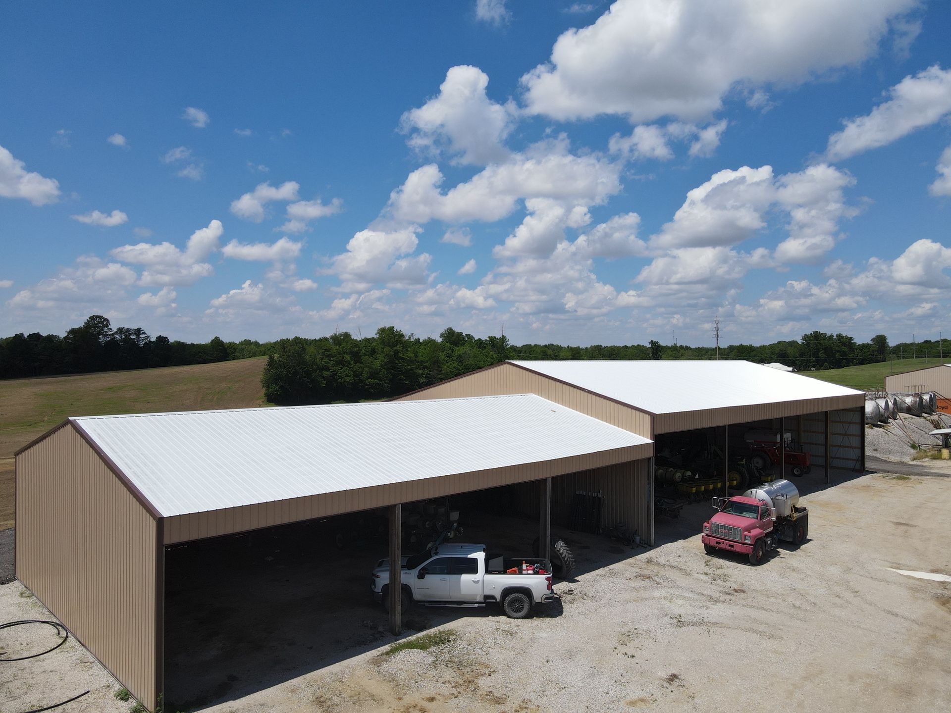 Open-sided tan metal carport with white roofs, sheltering vehicles in a gravel lot under a partly cloudy sky