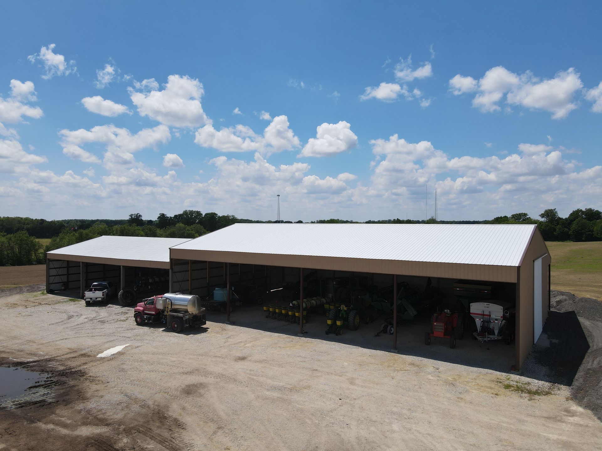 Rural metal barn with open bays, parked tractor, and gravel lot under a blue sky with clouds