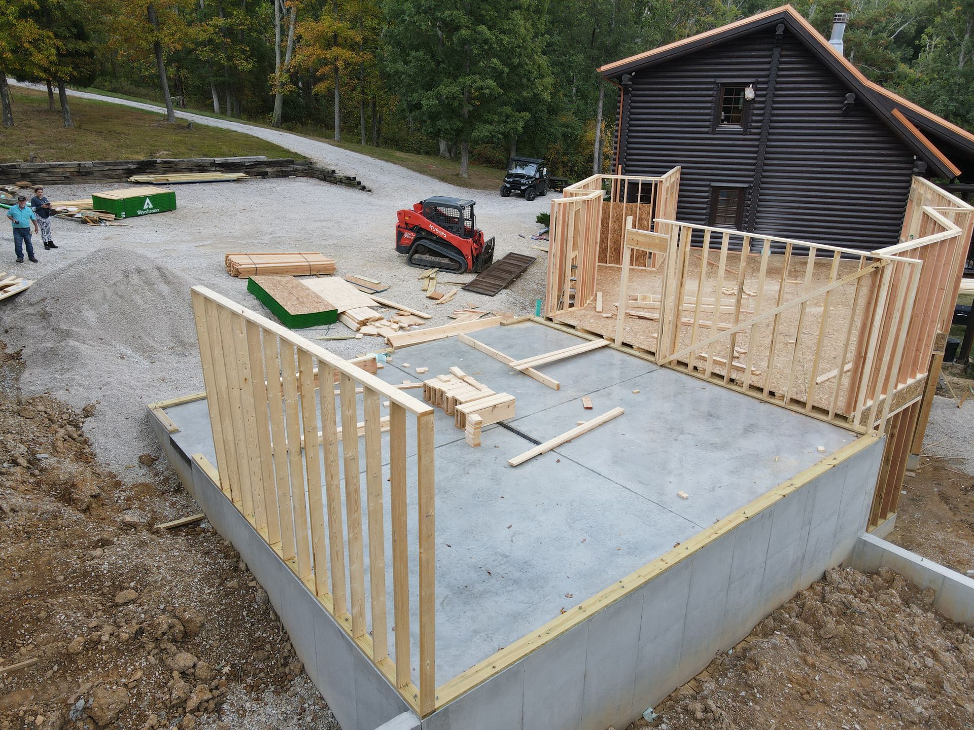 Concrete foundation with framed wooden walls under construction beside a cabin and gravel driveway.