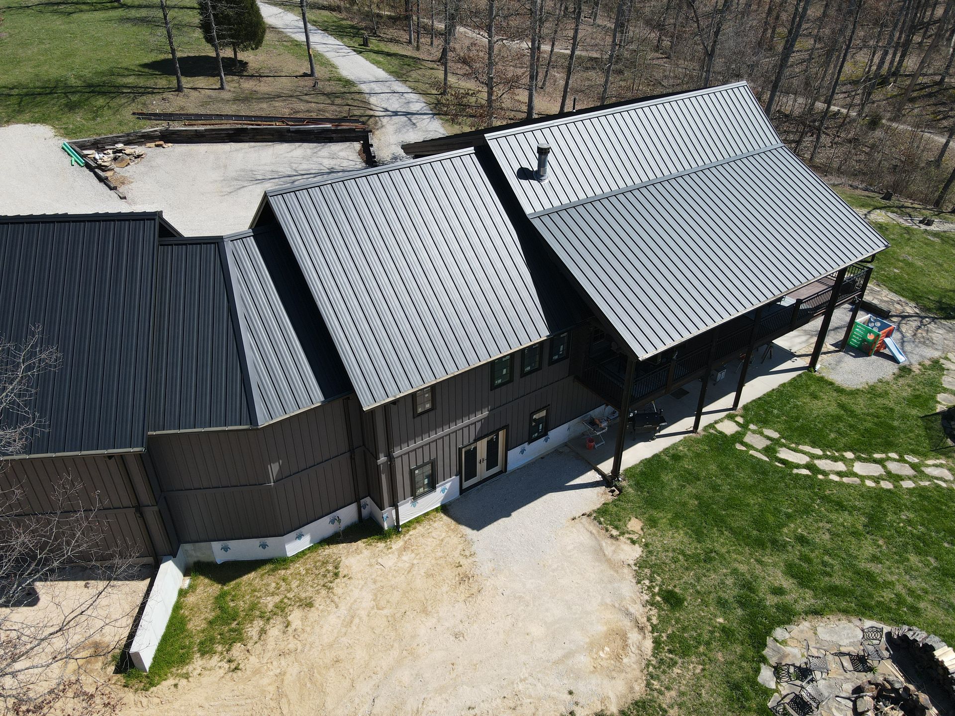 Aerial view of a dark-roofed house with a porch, gravel yard, and grassy lawn.