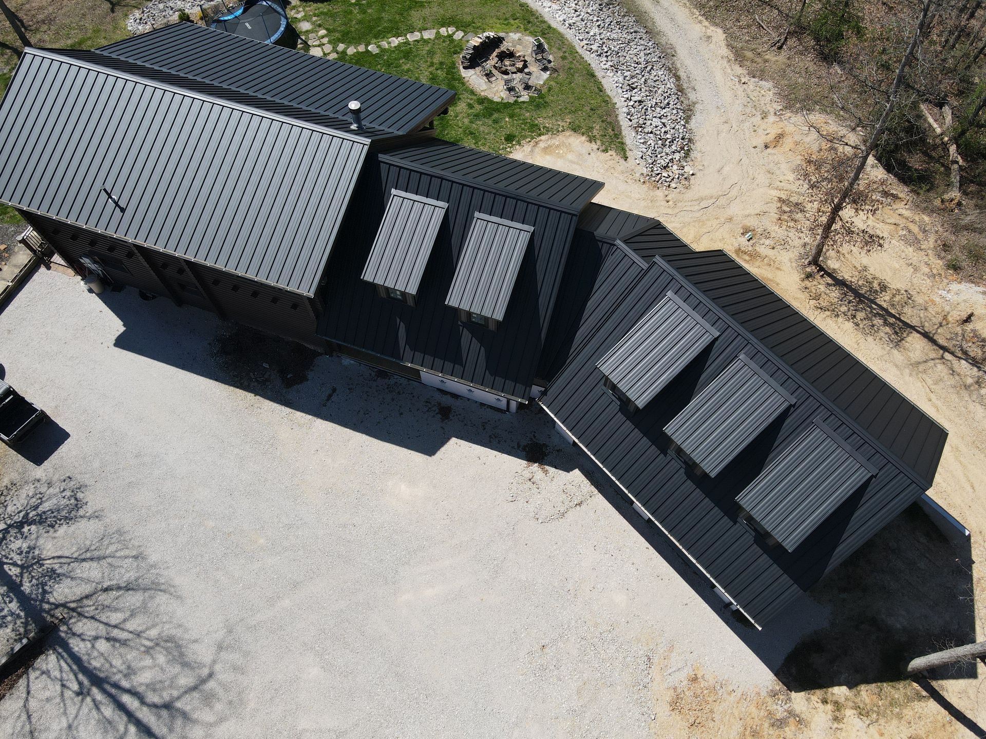 Aerial view of a black-roofed modular building beside a gravel driveway and wooded area