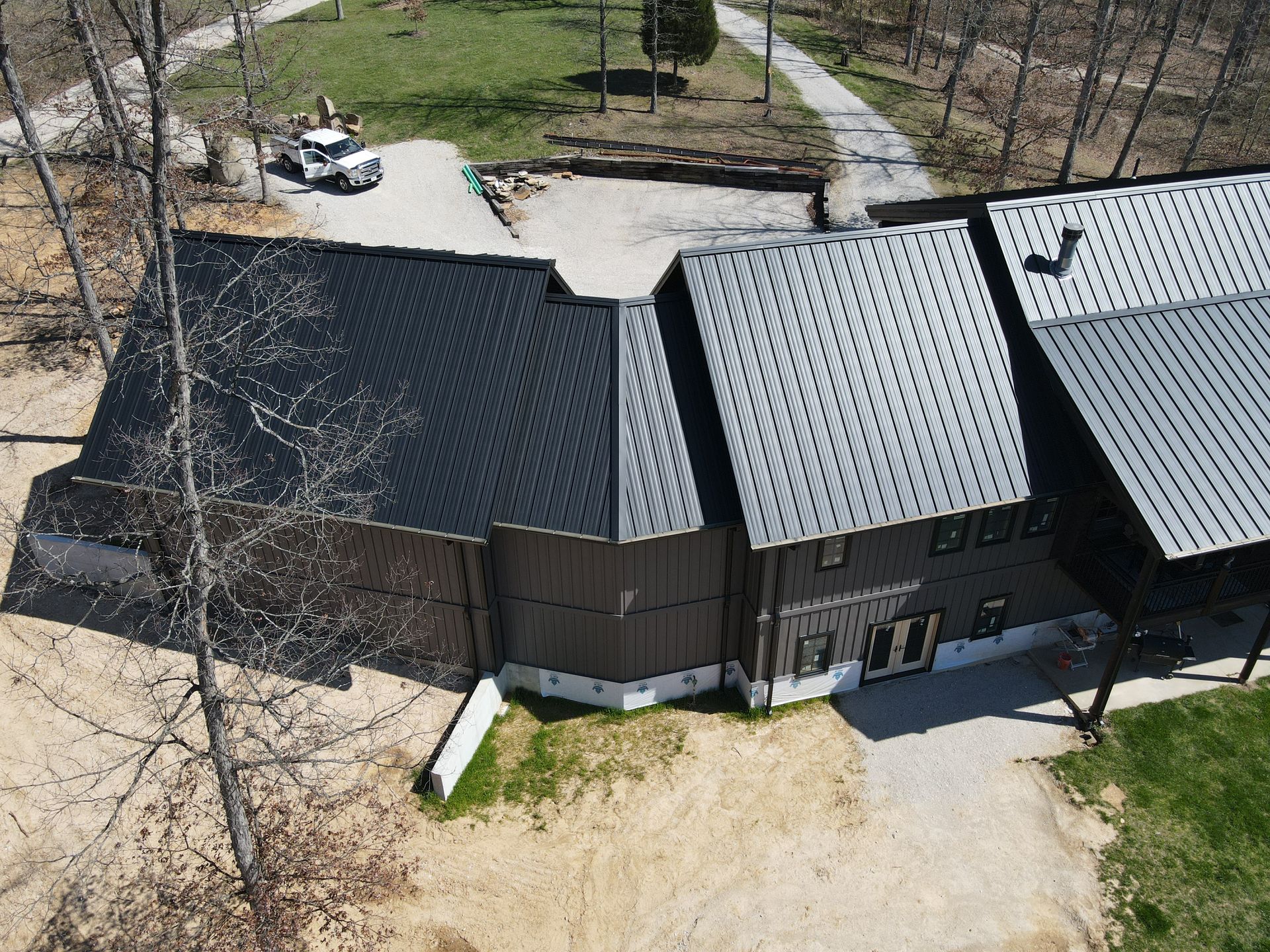 Aerial view of a modern house with dark and gray roofs, surrounded by trees and a dirt yard.