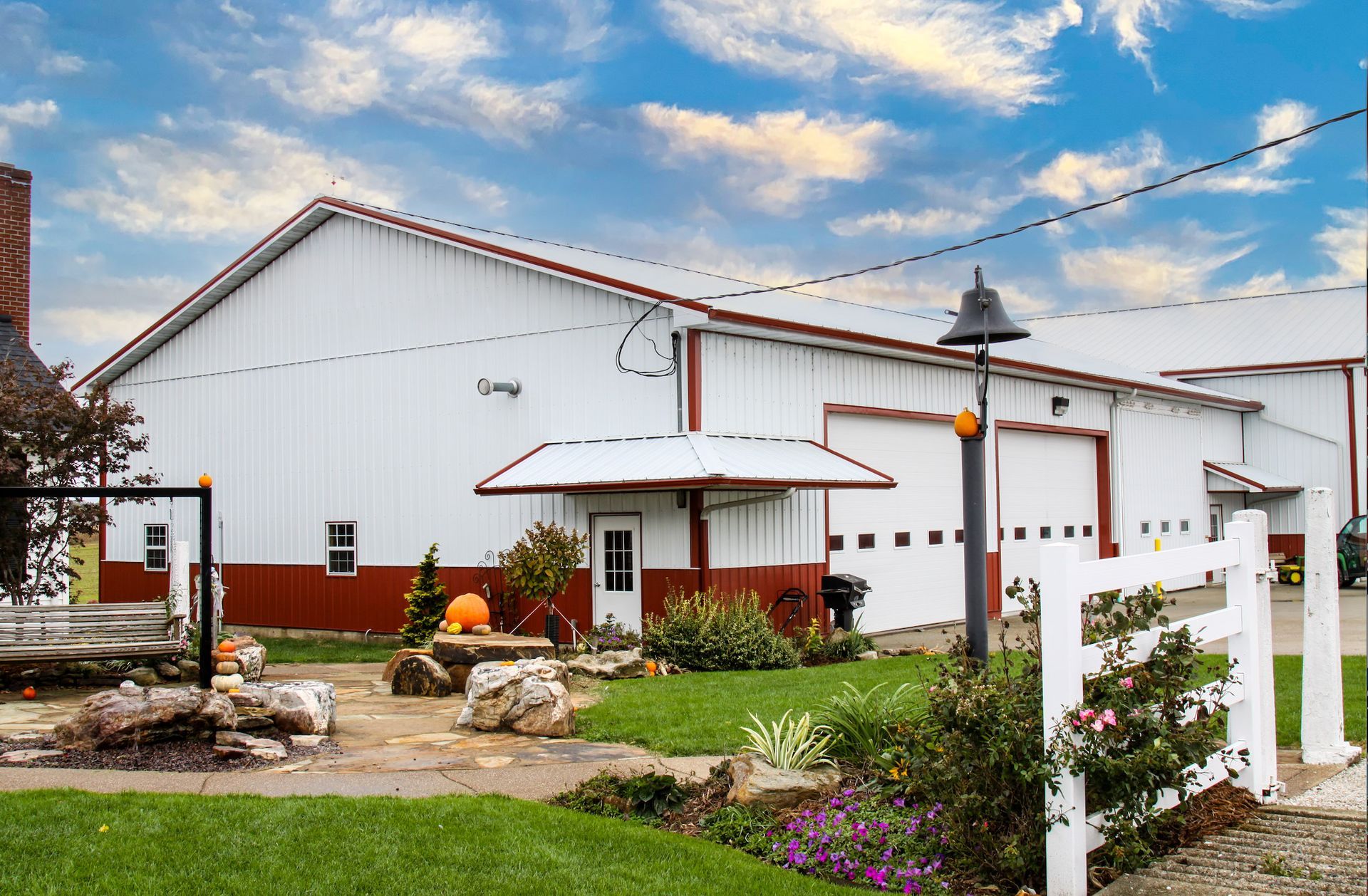 White barn-style building with red trim, a small garden, and a white fence under a blue sky
